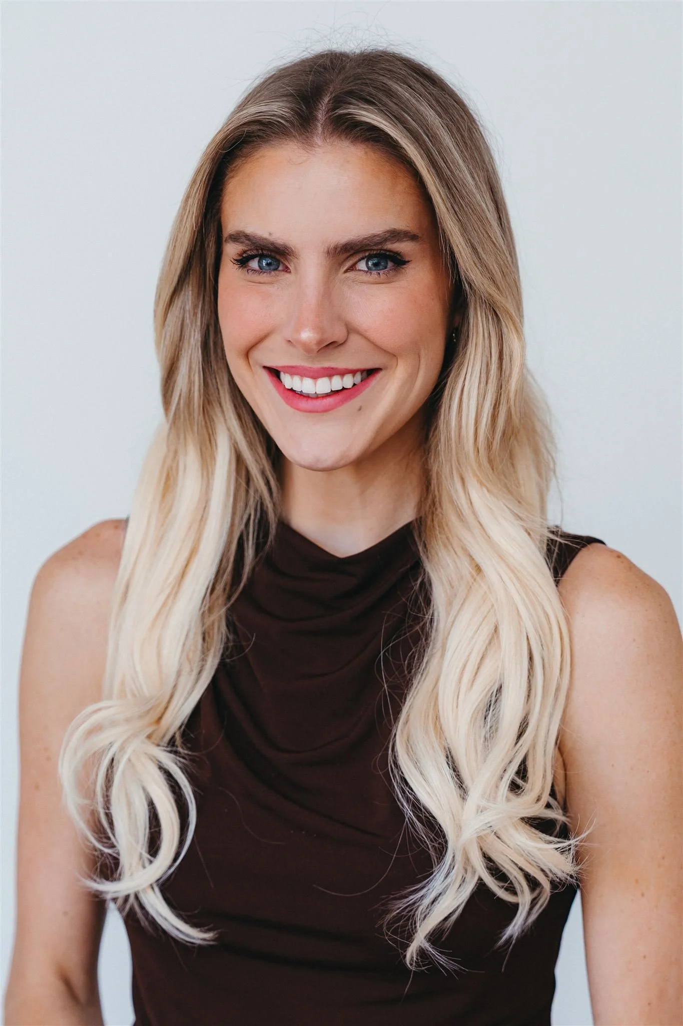 A woman with long blonde hair, blue eyes, and a bright smile, wearing a sleeveless dark brown top, standing against a plain white background.