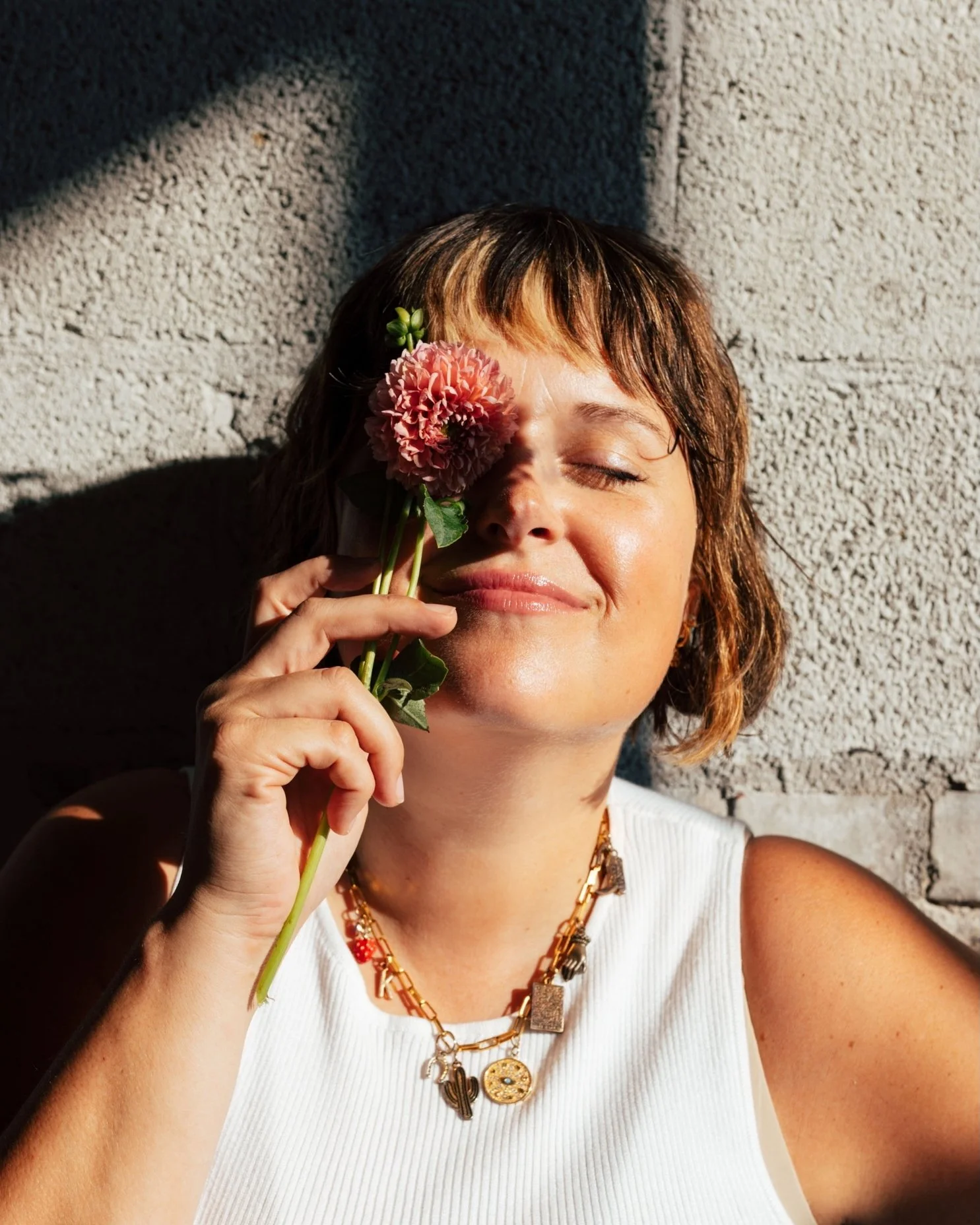 A woman with short brown hair and a gold chain necklace is holding a pink flower to her face, with her eyes closed and a content smile, standing against a textured gray wall.