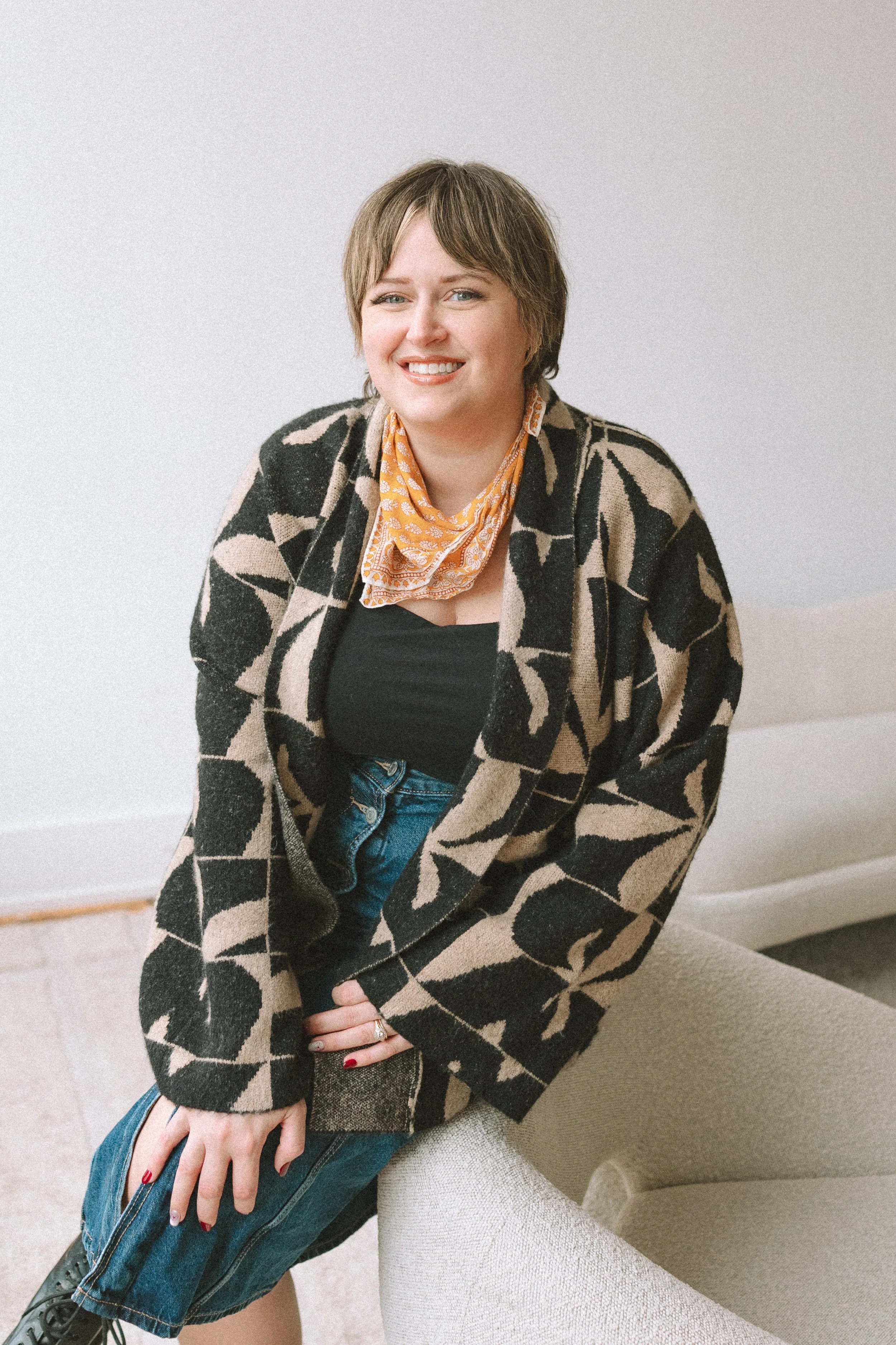 A woman with short brown hair, smiling, wearing a black top, patterned jacket, blue jeans, sitting on a beige chair in a bright room.