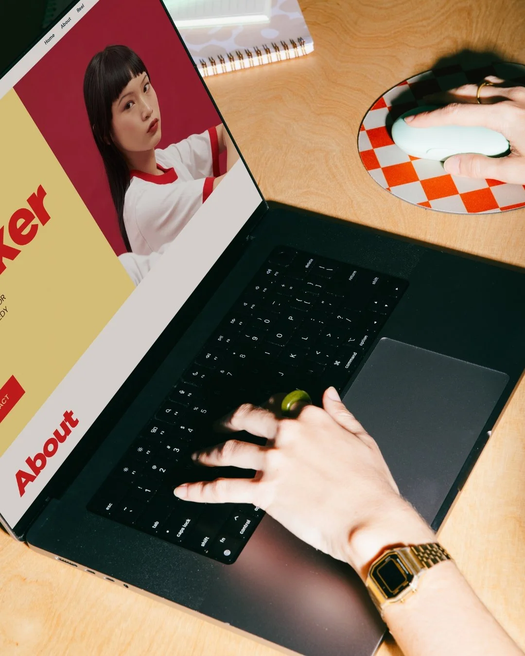 Person using a MacBook laptop with a website page open and a hand on the keyboard, sitting at a wooden desk with a mouse on a patterned mouse pad, a spiral notebook in the background.