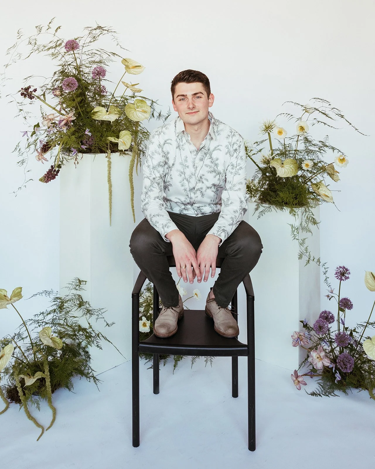 A young man sitting on a black chair surrounded by white backdrop and floral arrangements with green, purple, and yellow flowers and foliage.