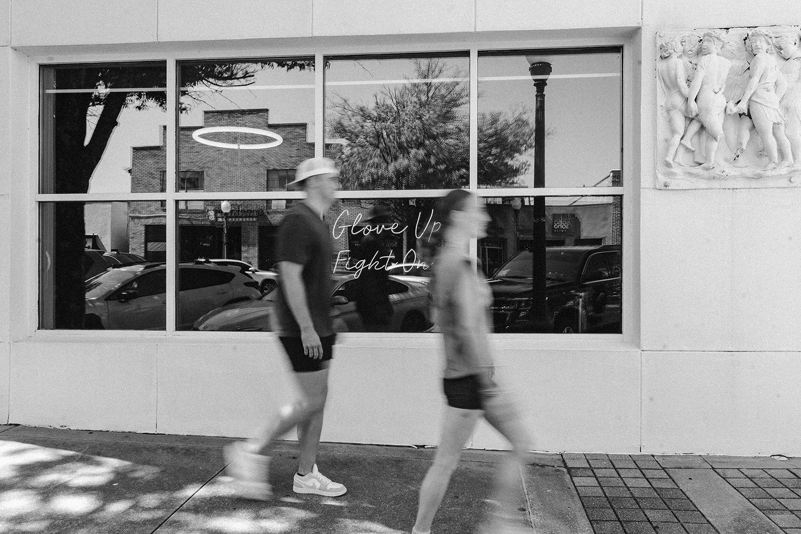 Two blurred people walk past a large window with a neon sign that says 'GloVe Up Fight On.' The window reflects parked cars, a brick building, trees, and a lamppost. There is a decorative stone relief sculpture on the building's wall.