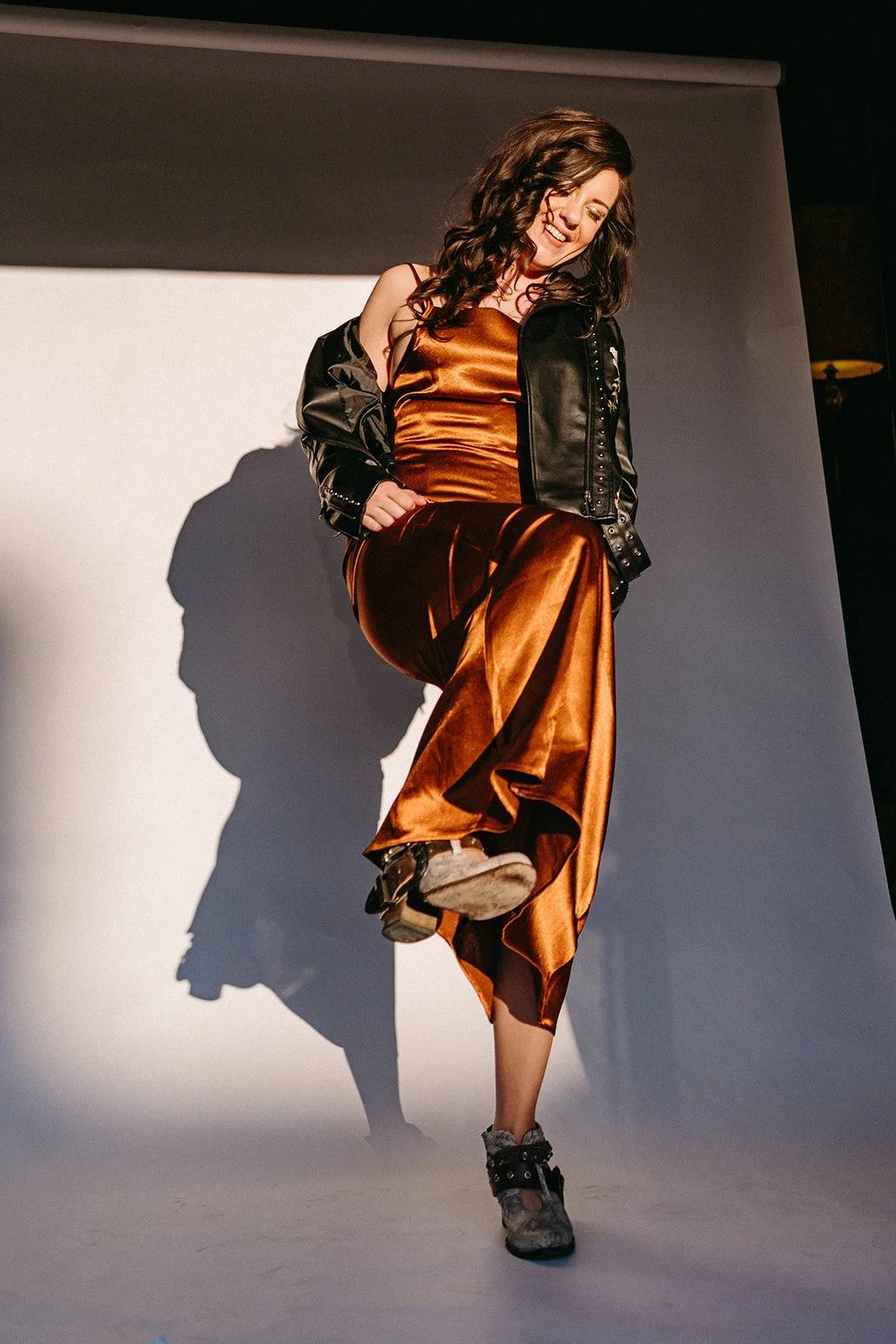 A woman posing and smiling in a photoshoot studio, wearing a shiny bronze dress, a black leather jacket, and sneakers. She is lifting one foot, creating a shadow on the backdrop.