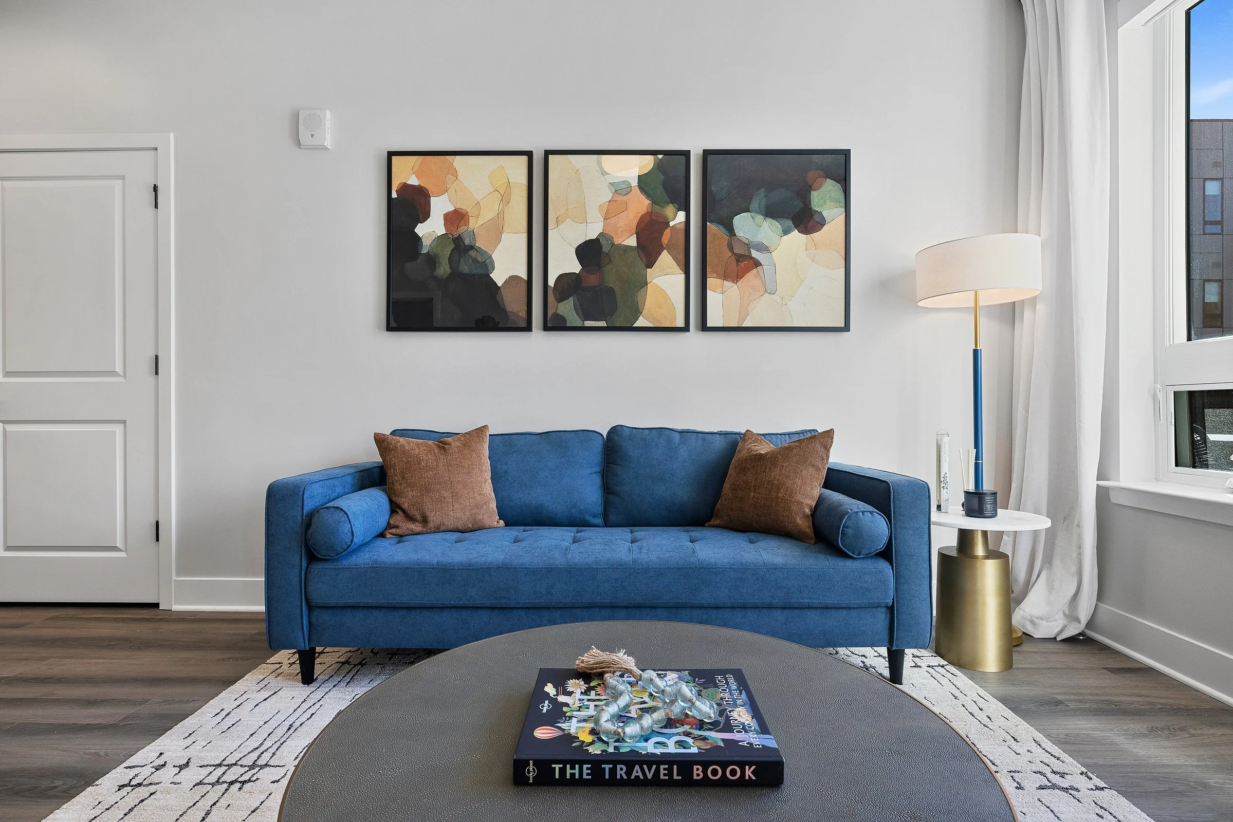 Modern living room with a blue sofa, brown pillows, abstract art on white wall, white curtains, floor lamp with white shade, window with view outside, and a coffee table with a travel book and decor on top.