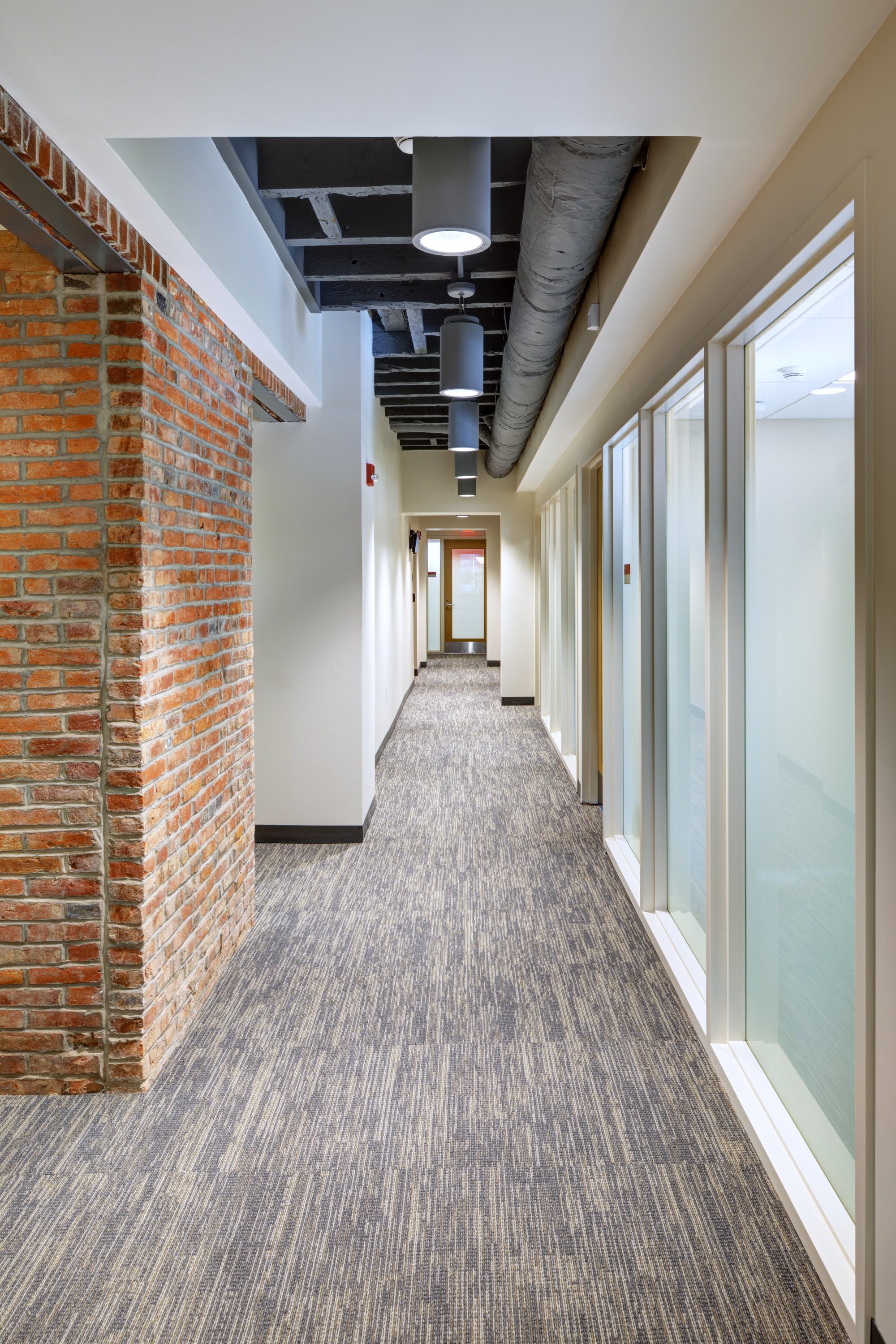 Empty office hallway with brick wall on the left, white walls on the right with frosted glass windows, carpeted floor, and exposed ceiling with black ducts and hanging cylindrical light fixtures.