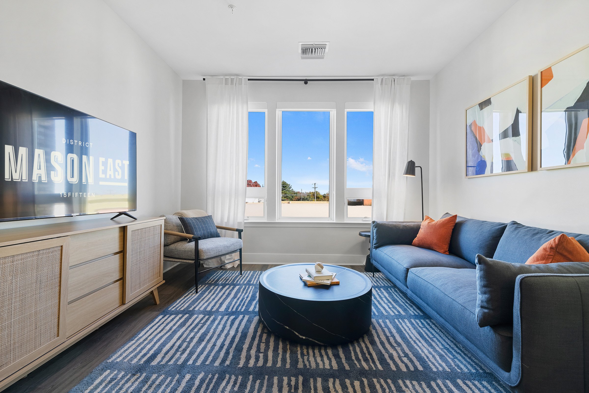 Bright living room with white walls, large window with white curtains, blue sofa with orange and gray pillows, abstract art on the wall, round coffee table with books and decorative items, patterned rug, armchair, floor lamp, and flat-screen TV.