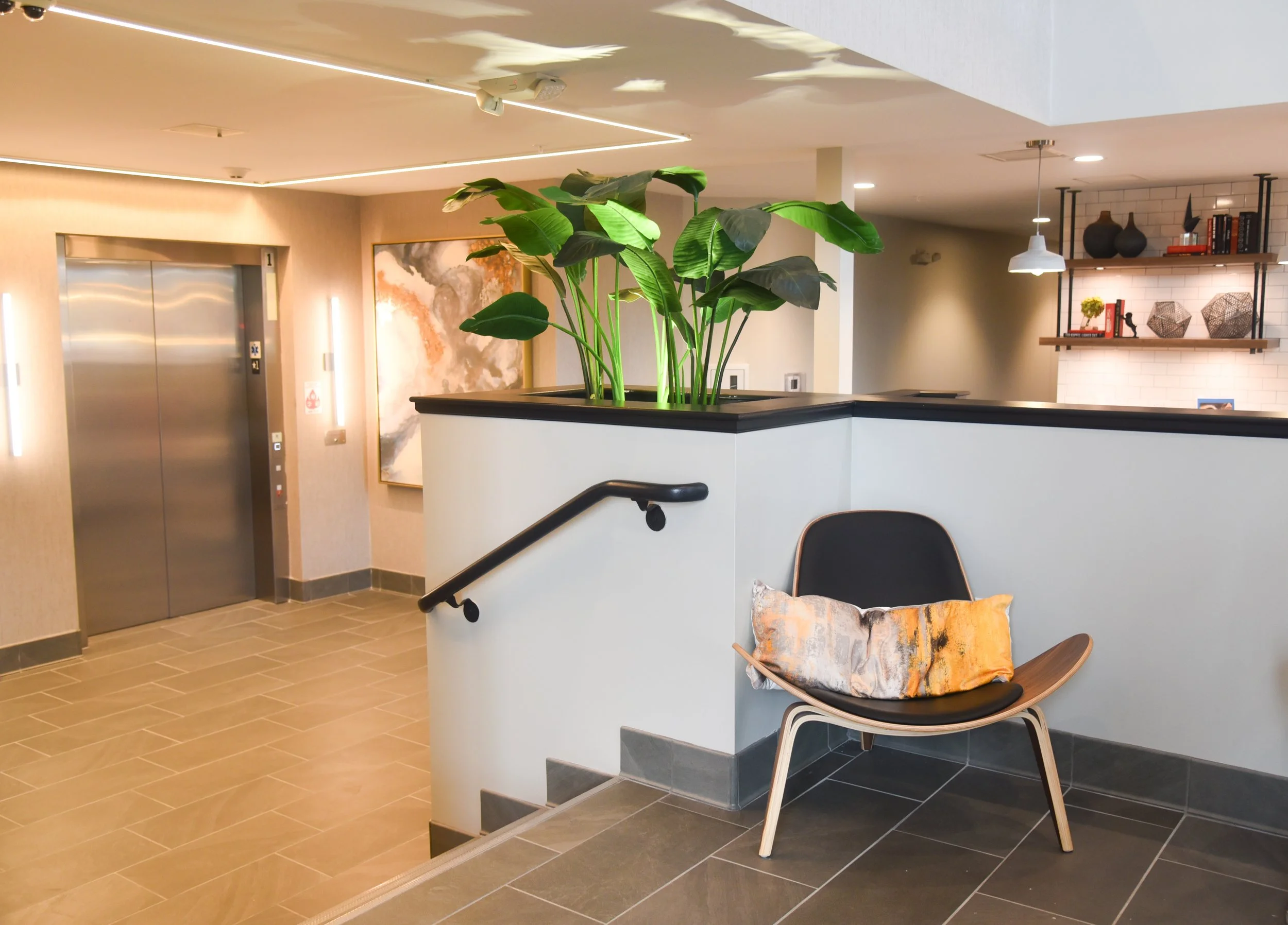 Modern interior space with an elevator, a potted plant on a partition, and a stylish chair with a decorative pillow in front.