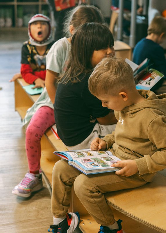 Children sitting on a wooden bench, reading books in a library with colorful shelves in the background.