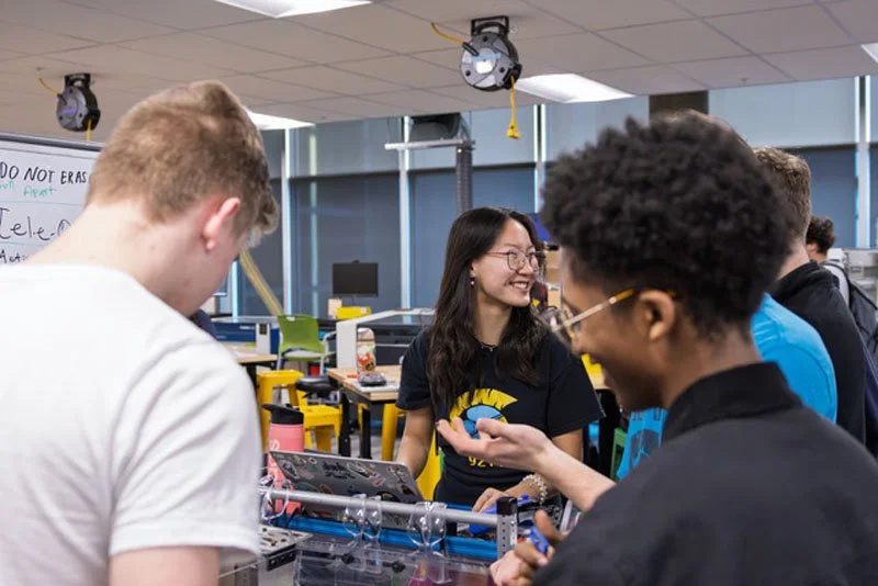 A group of students in a classroom engaged in a robotics project, with one girl smiling and talking to two boys.