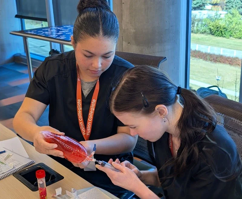 Two women in black scrubs with lanyards that say 'Fusion Collegiate' are in a room with large windows. One woman is holding a large syringe and a blood bag, while the other is preparing to draw blood from her arm.