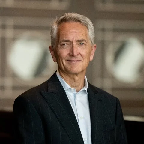 Portrait of an older man with gray hair in a business suit, smiling in an indoor setting.