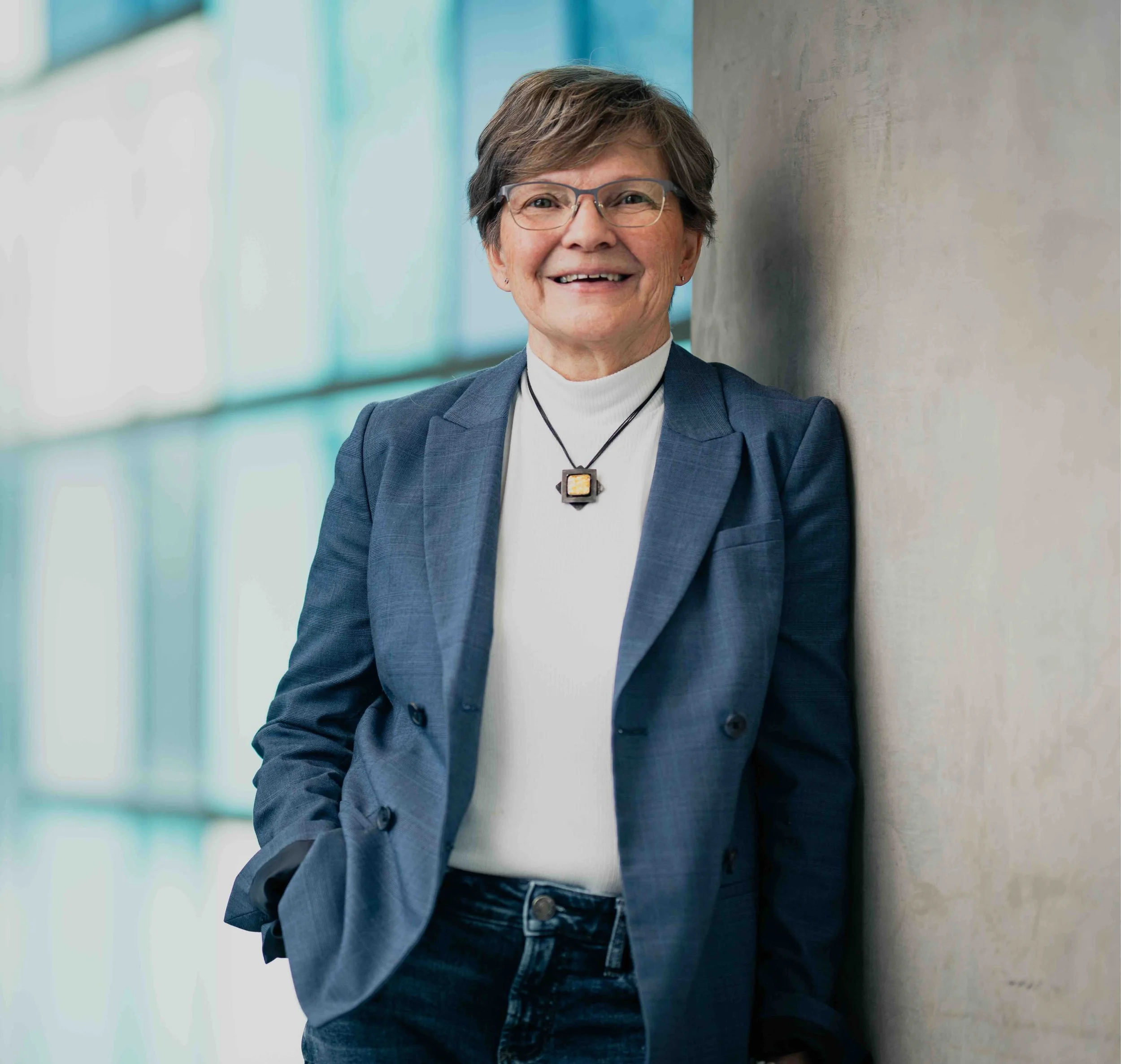 Portrait of a smiling woman with short gray hair, glasses, wearing a white turtleneck, blue blazer, and necklace, leaning against a beige wall in a modern building with large windows.