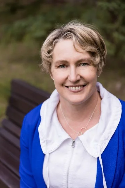 A woman with short curly blonde hair, wearing a white hoodie and blue jacket, smiling outdoors with trees and a park bench in the background.