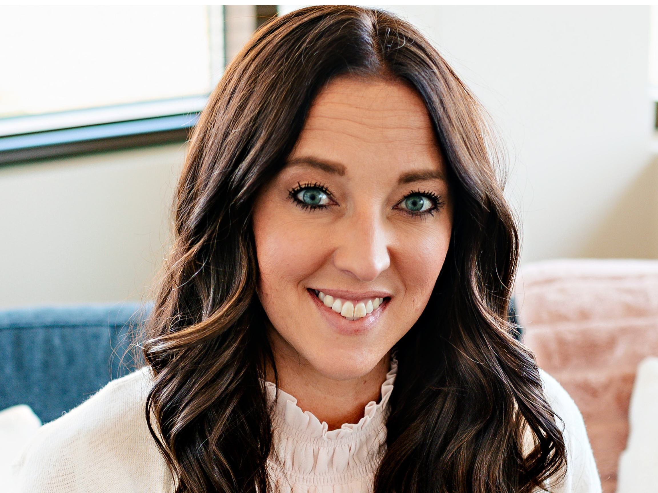 Close-up of a woman with long, wavy brown hair, smiling, sitting indoors near a window with blinds.