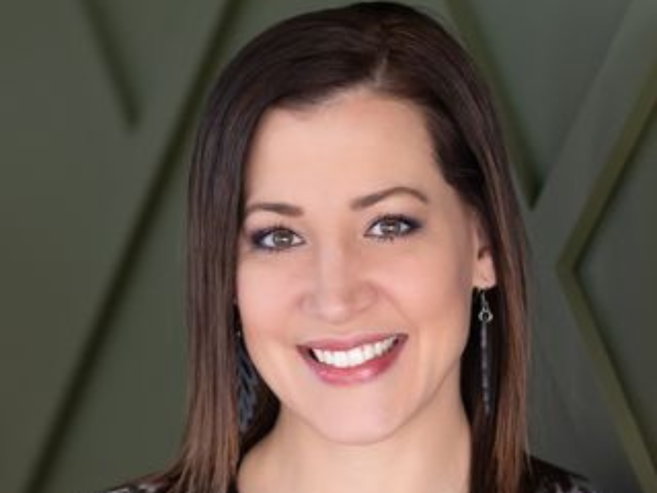 Close-up of a woman with long brown hair, smiling, wearing earrings, against a green background.