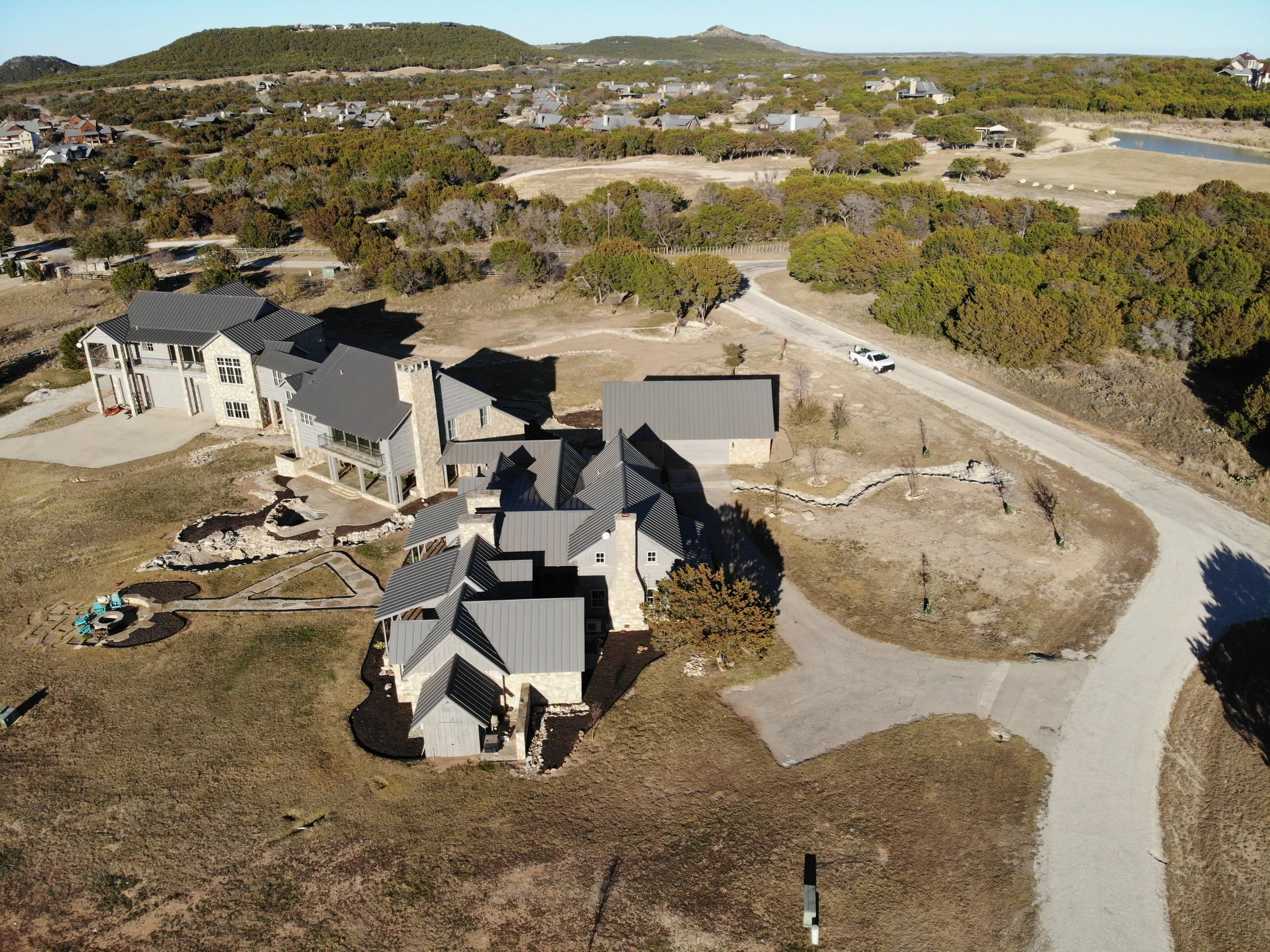 Aerial view of a large modern house with gray metal roof in a rural area, surrounded by a dirt road, trees, and an open landscape with hills in the background.