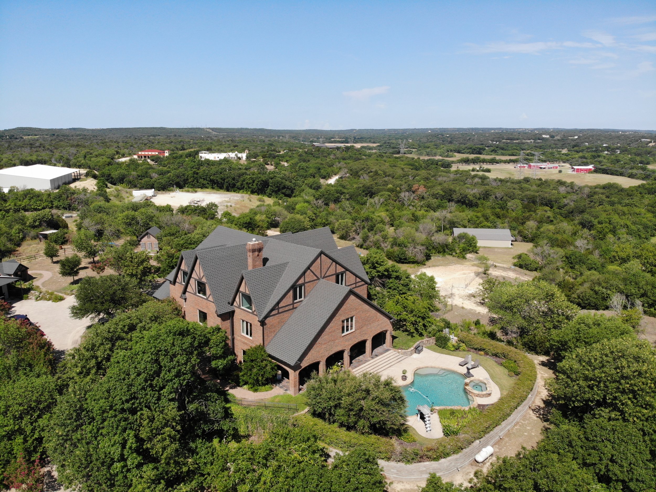 Aerial view of a large brick house with a steep gray roof, surrounded by lush green trees, with a swimming pool and hot tub in the backyard.