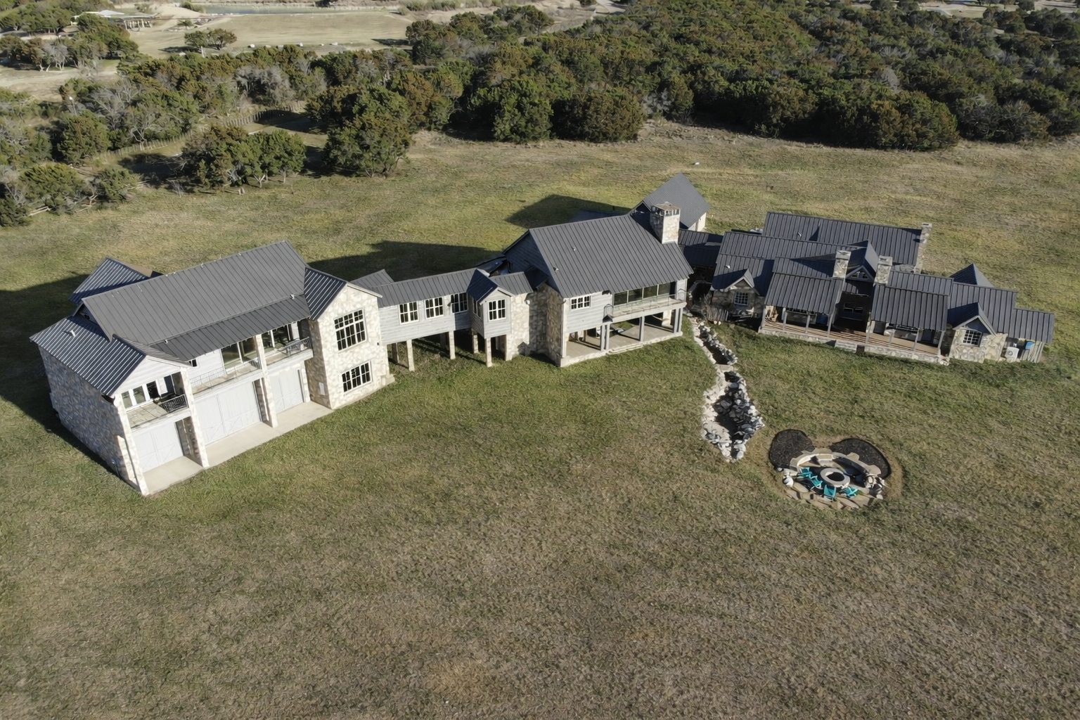 A large modern house with a dark gray metal roof, stone and wooden exterior, and multiple sections on a spacious grassy lot with a decorative fire pit and landscaping.