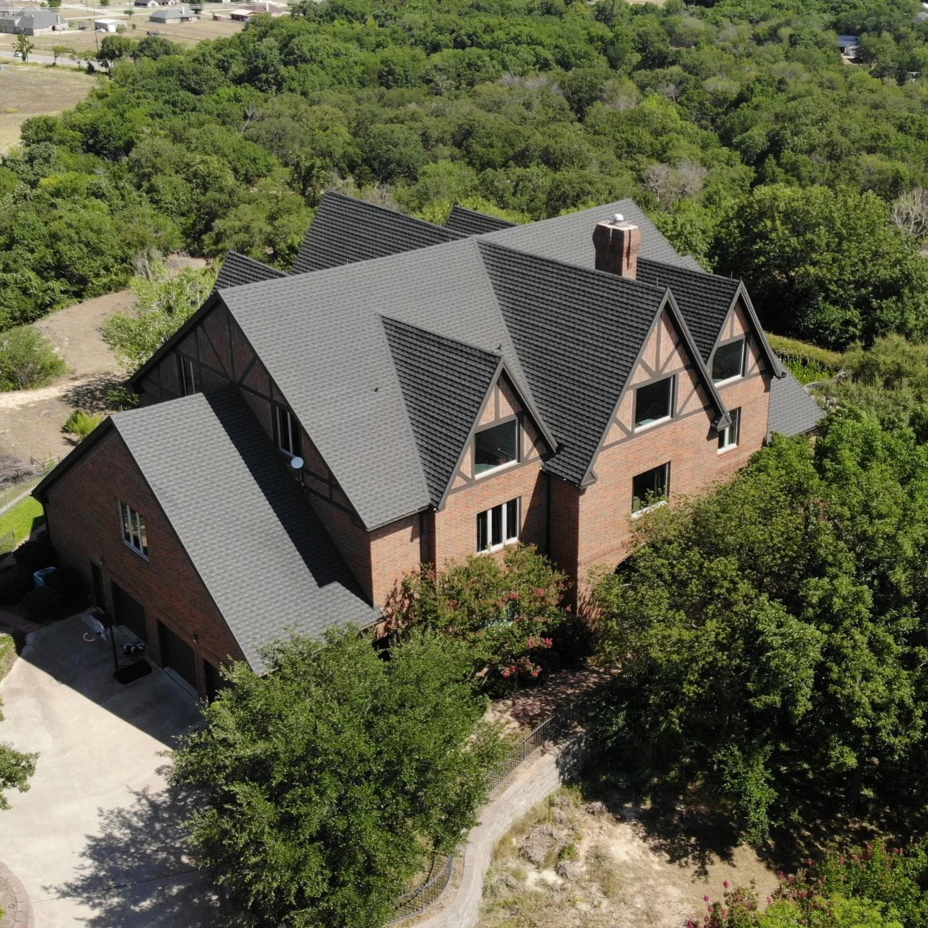 An aerial view of a large brick house with a complex dark gray tiled roof surrounded by trees and greenery.