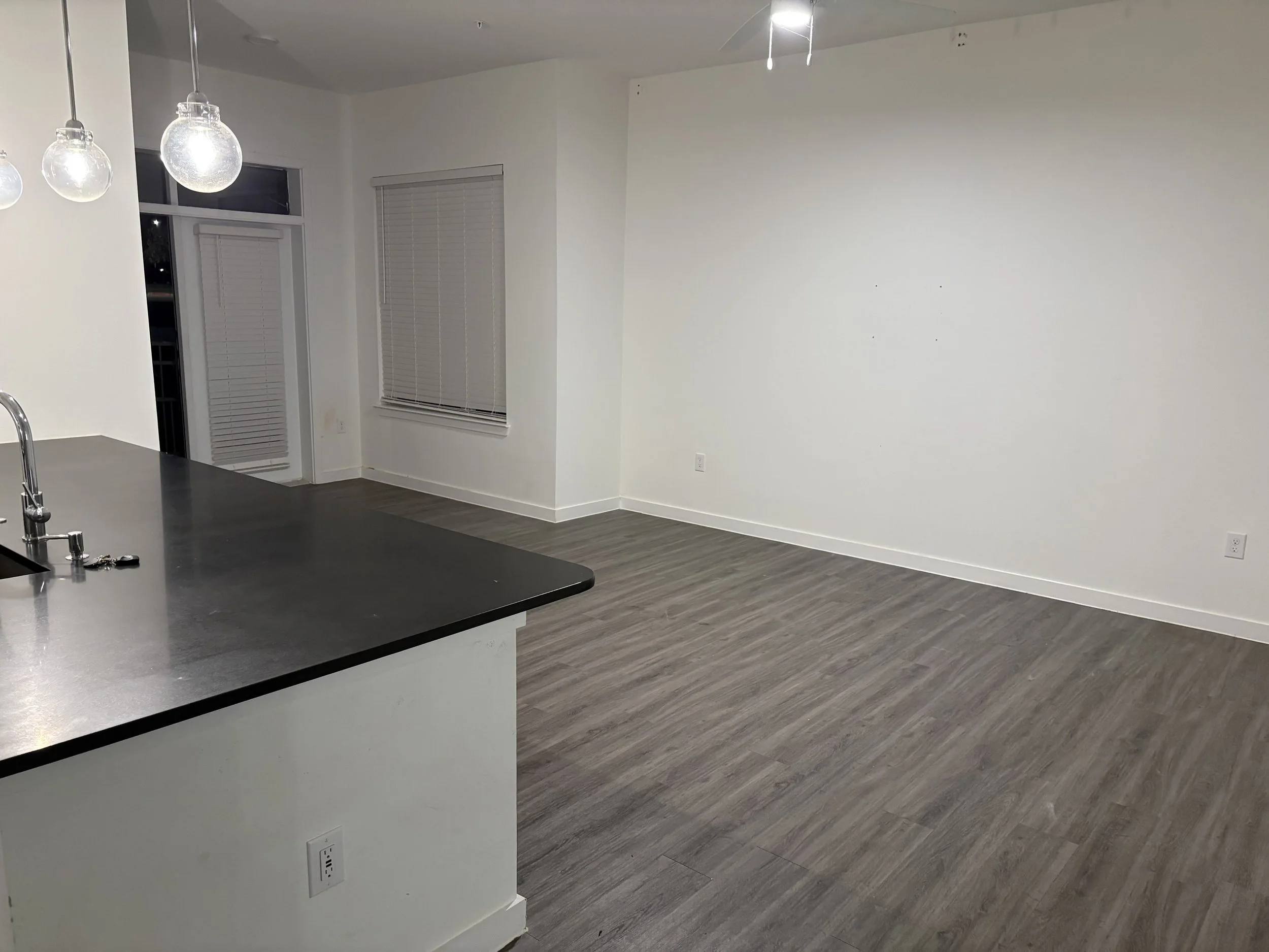 Empty living room with dark wood flooring, white walls, and windows with closed blinds. Corner of a kitchen island with a black countertop and a sink with a faucet is visible, with a key on the counter.