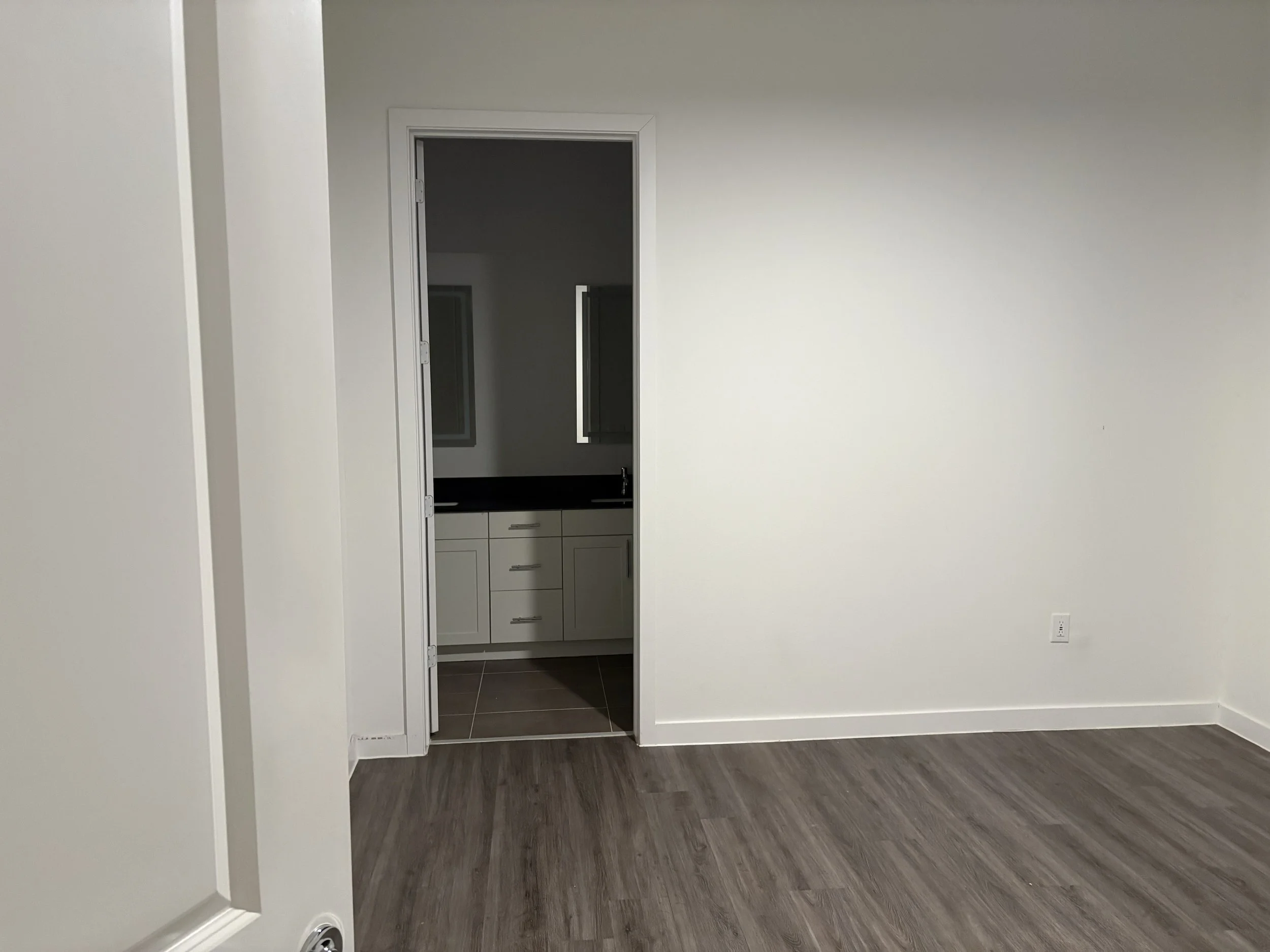 An empty room with white walls, a wooden floor, and a doorway leading to a small room with a sink and mirror.