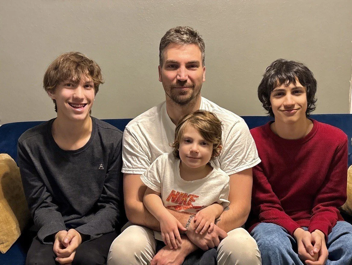 A man with three boys sitting on a dark blue couch against a beige wall.