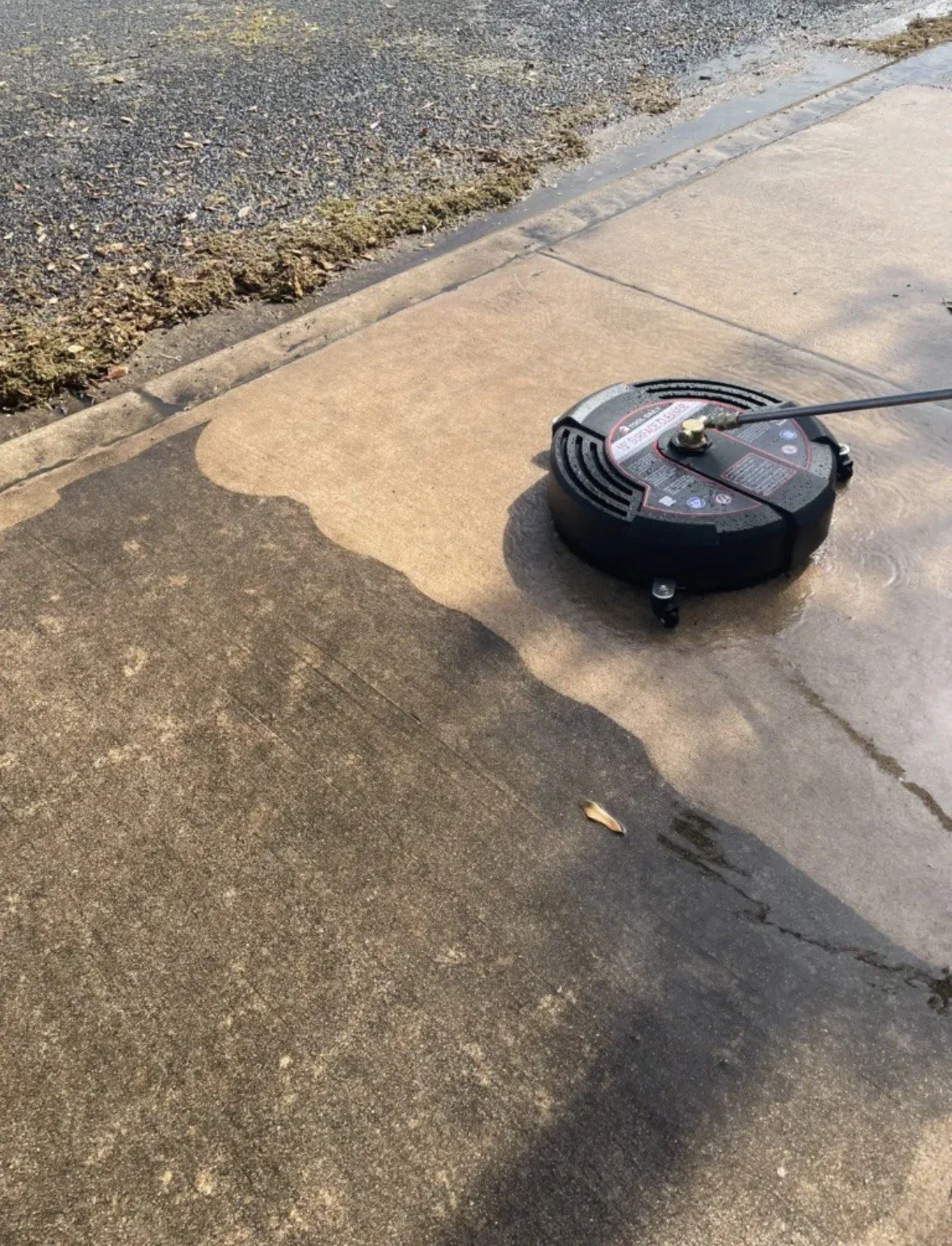 A concrete sidewalk with a round, black pressure washer on it. Water is spraying from the pressure washer onto the sidewalk, cleaning part of the surface. The area being cleaned is visibly lighter in color, contrasting with the dirtier, darker sectio