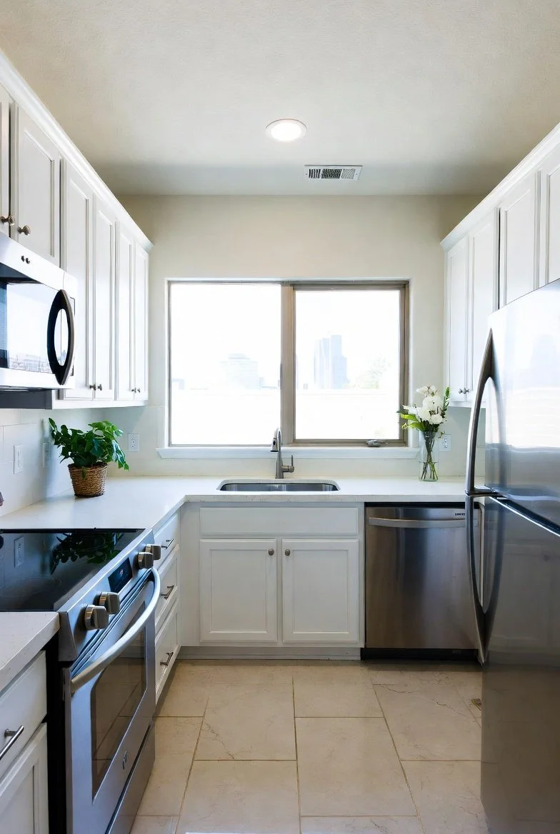 A bright, modern kitchen with white cabinets, a stainless steel refrigerator, stove, and dishwasher, a window above the sink, and decorative plants on the counter.