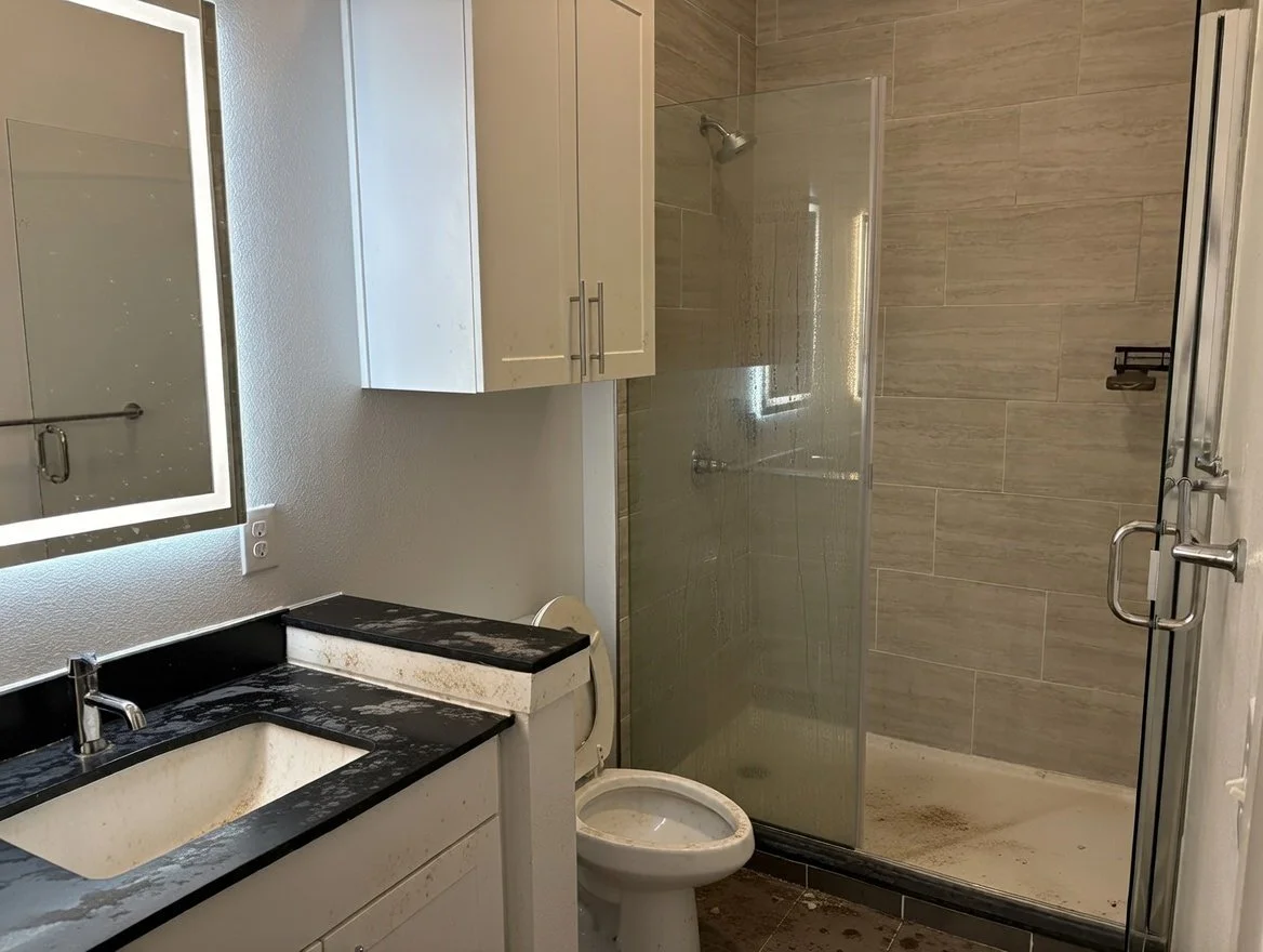 A bathroom with a black and white marble countertop sink, a mirror with built-in lighting, a white cabinet, a toilet, and a glass-enclosed shower with beige tile walls and a metal handle on the door. The shower floor has dirt or stains.