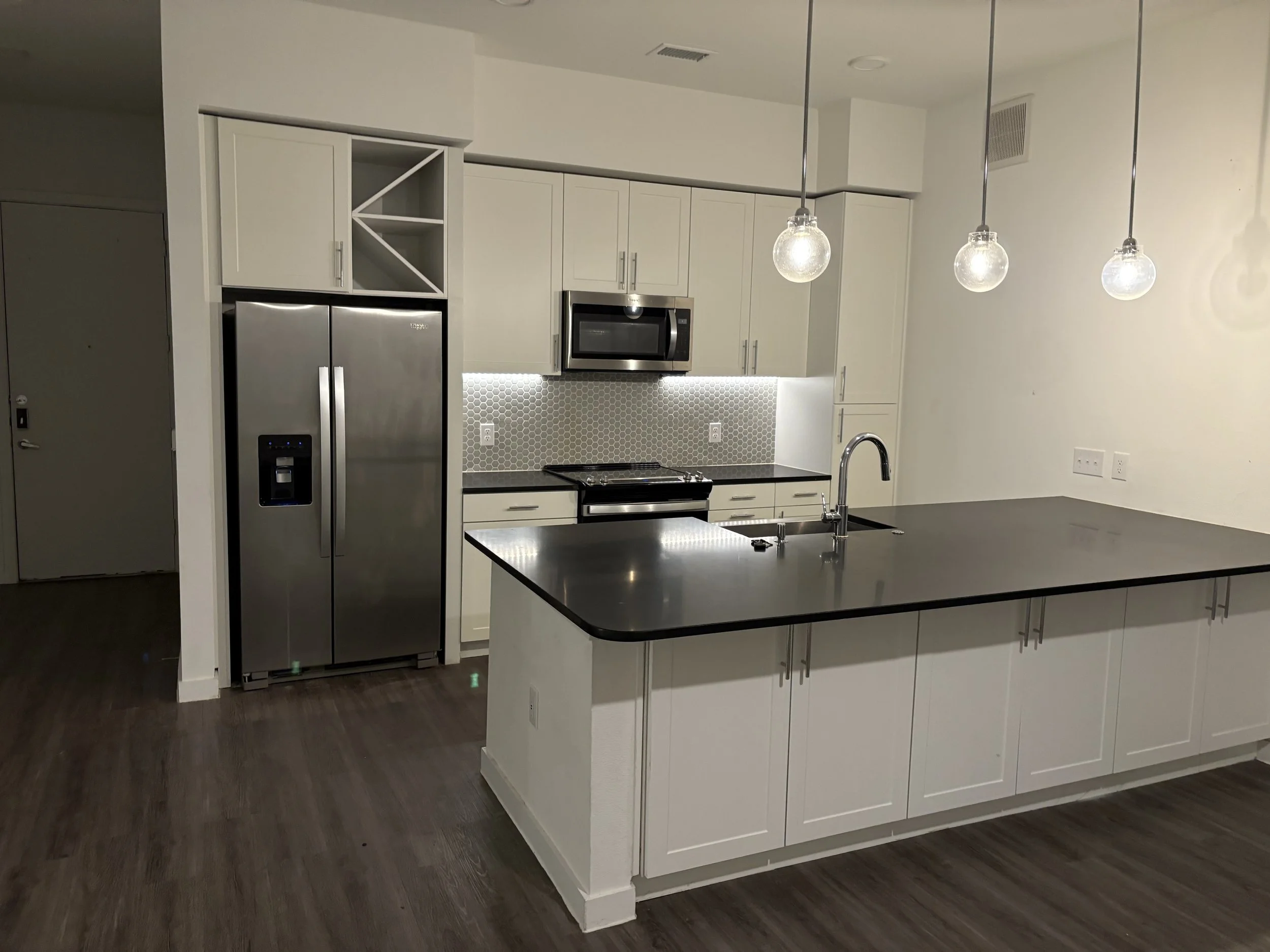 Modern kitchen with white cabinets, black countertop island, stainless steel refrigerator and microwave, small hexagon tile backsplash, and three hanging pendant lights.