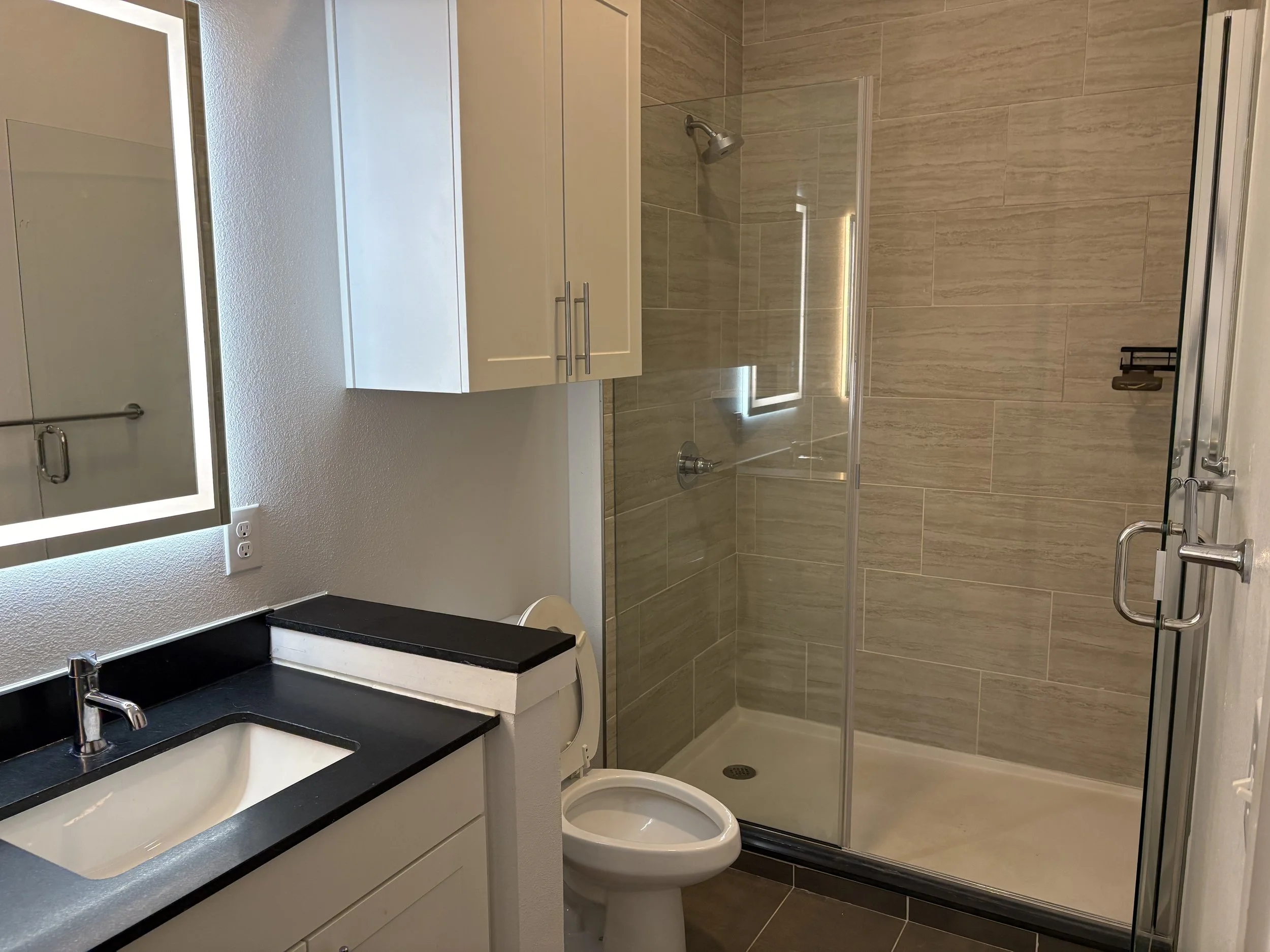 Bathroom with a black countertop sink, white cabinets, a wall-mounted mirror with LED lighting, a toilet, and a walk-in shower with beige tiles and a glass door.