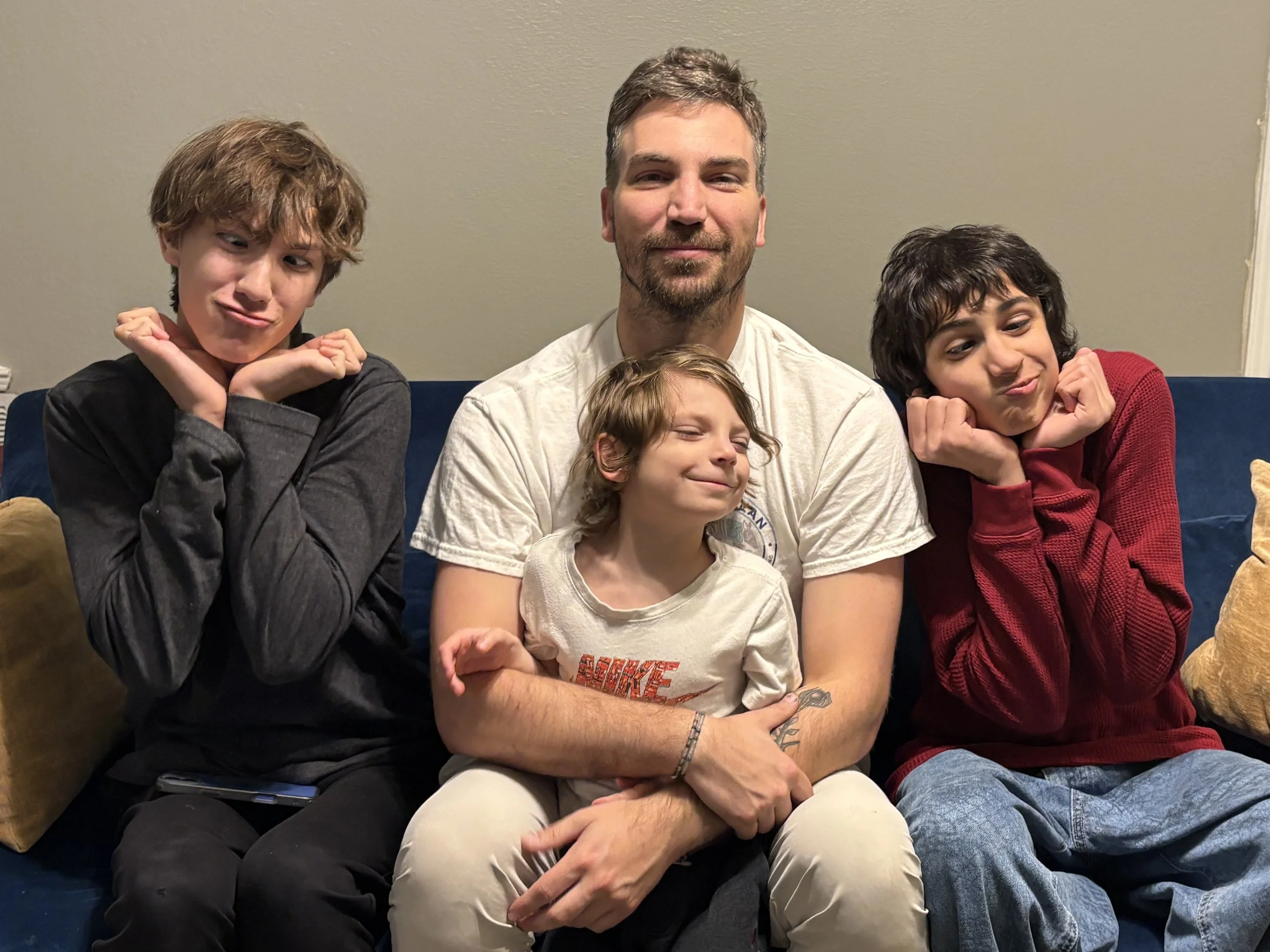 A man sitting on a blue couch with three children, all making playful, puckered expressions and posing with their hands under their chins.
