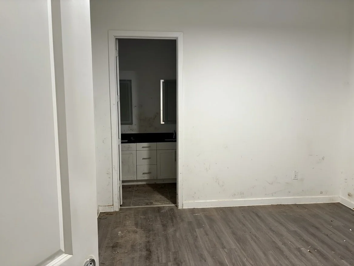 Empty room with dirty wooden floor, blank white wall, and open doorway leading to a kitchen with white cabinets and a black countertop.