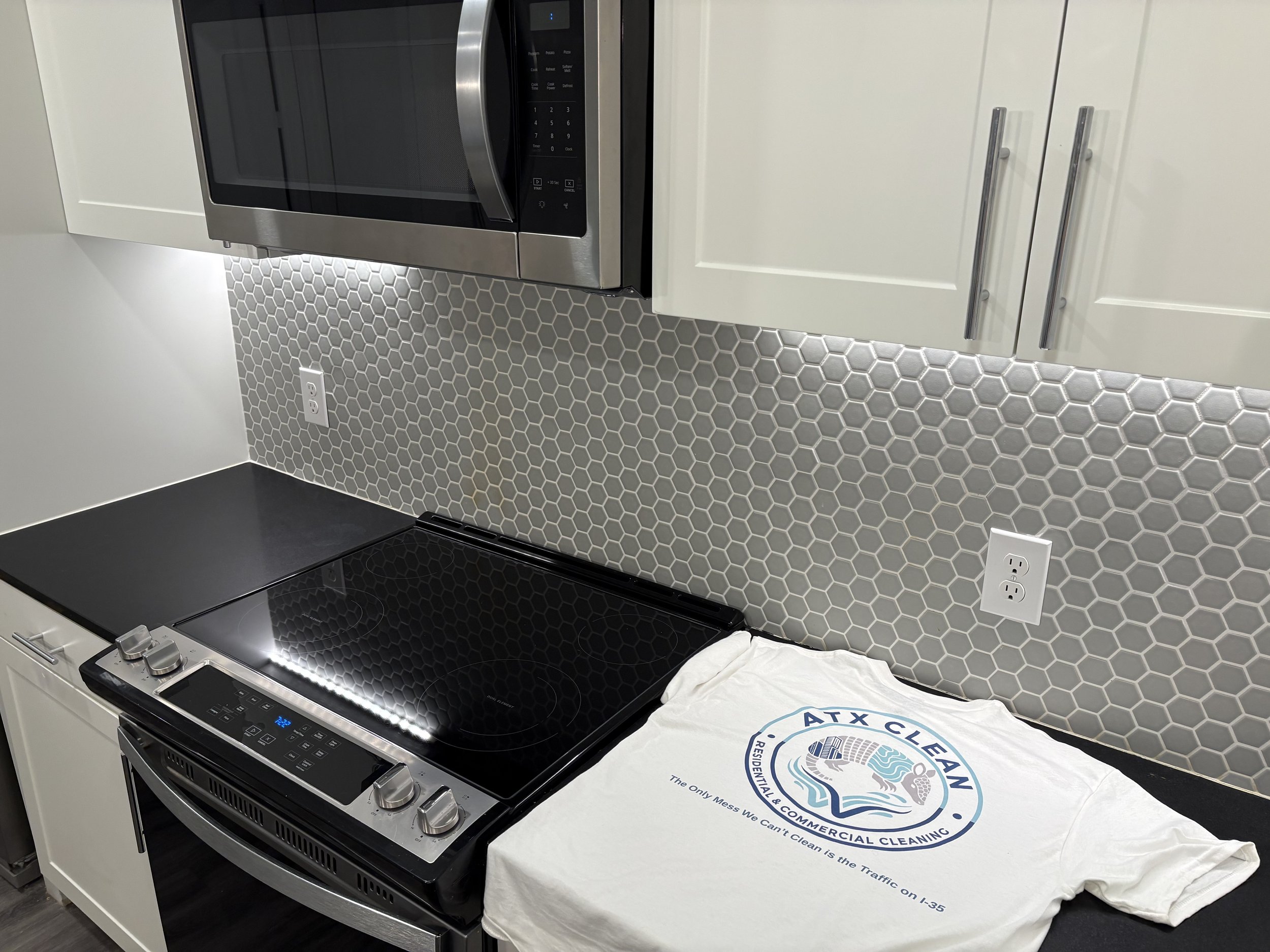 A modern kitchen with a black glass electric stove, a stainless steel microwave, white cabinets, a gray hexagonal tile backsplash, and a white t-shirt with a logo and text on it.