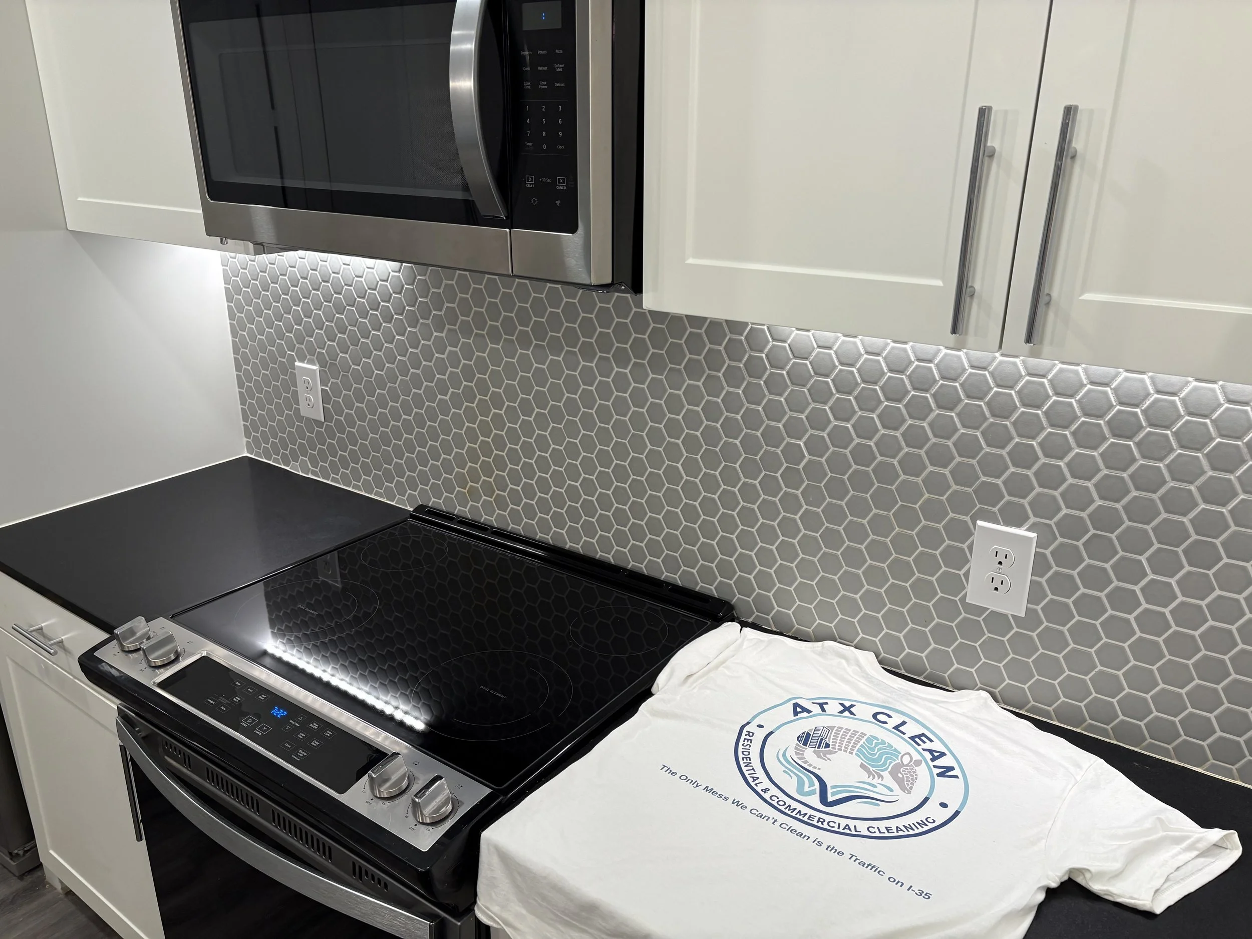 Kitchen countertop with a black induction stove, a white cabinet with silver handles, a microwave above, and a white cleaning t-shirt with a logo and text on it.