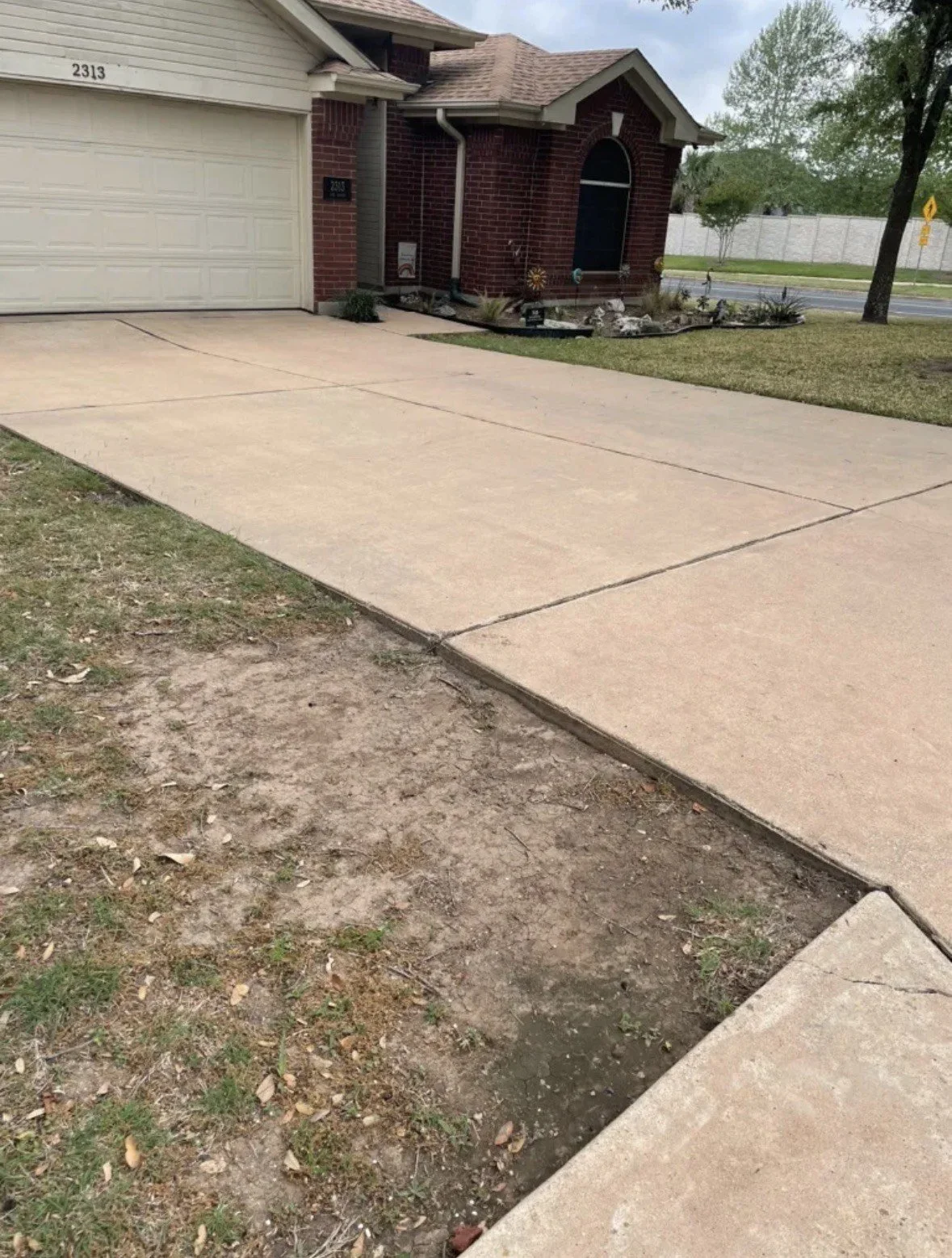 A suburban house with a brick exterior, a driveway, a garage, and a small front yard with a garden plot. The sidewalk is partially dug up, revealing dirt underneath.