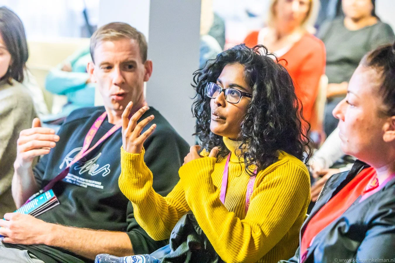 Een groep mensen zit in een zaal, een vrouw in een gele trui en een man in een zwarte trui lijken met elkaar te praten, anderen luisteren aandachtig.