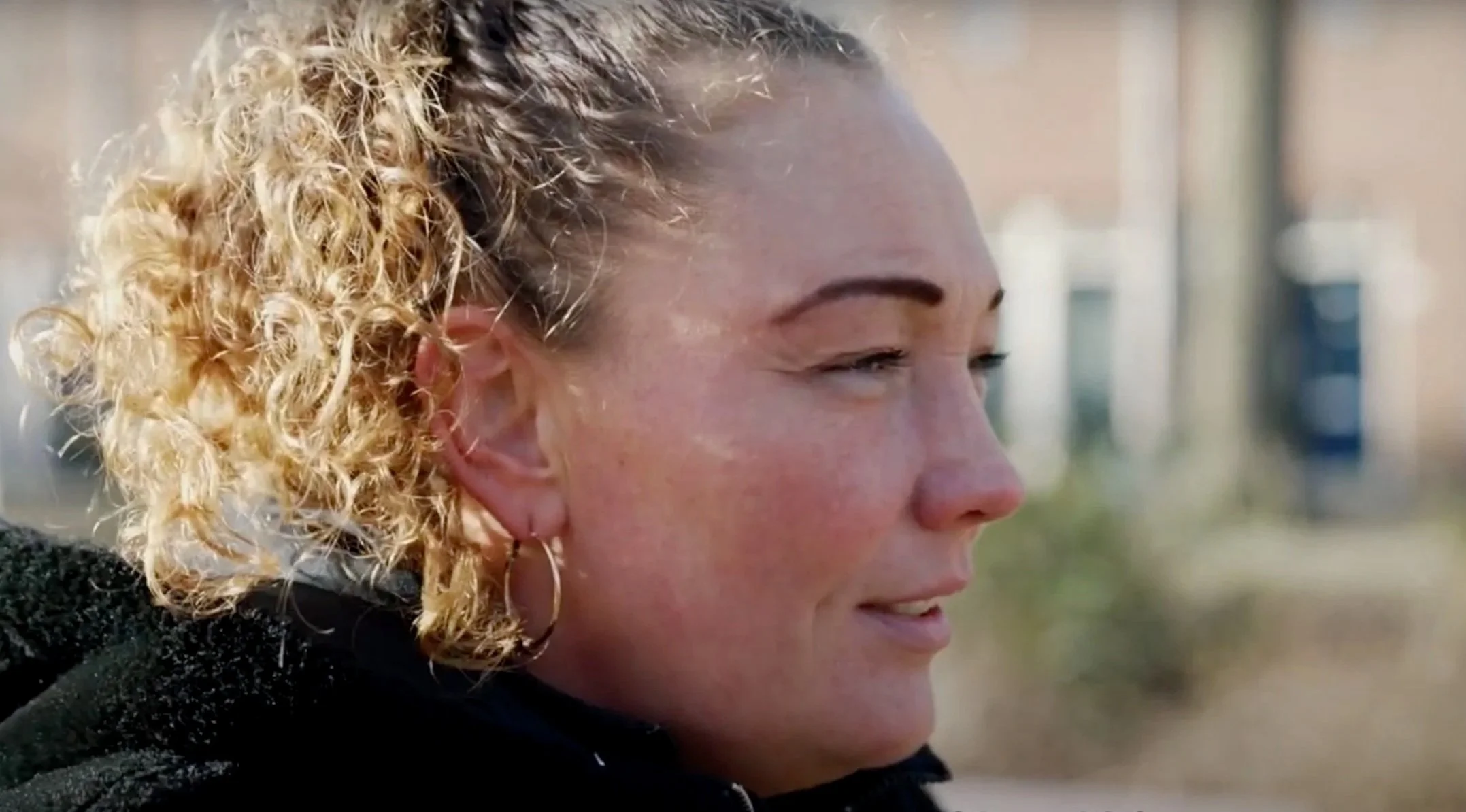 Close-up van een lachende vrouw met blond, gekruld haar, oorbellen en een zwarte jas, buiten met gebouwen op de achtergrond.