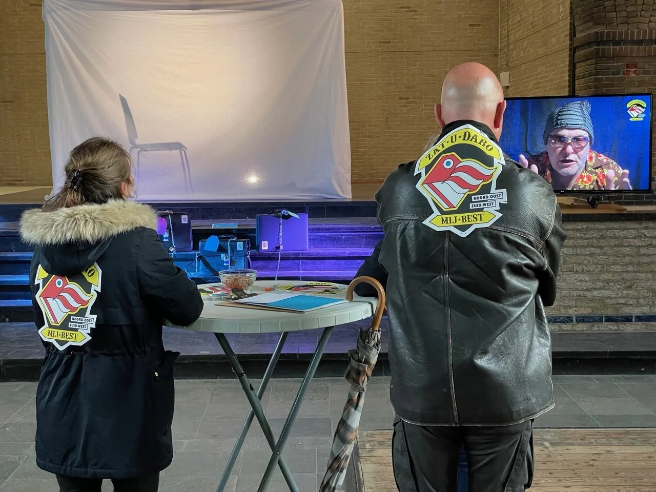 Twee mensen met opvallende badges kijken naar een tv-scherm waarop een man met een hoed en zonnebril spreekt, in een ruimte met brickwanden.