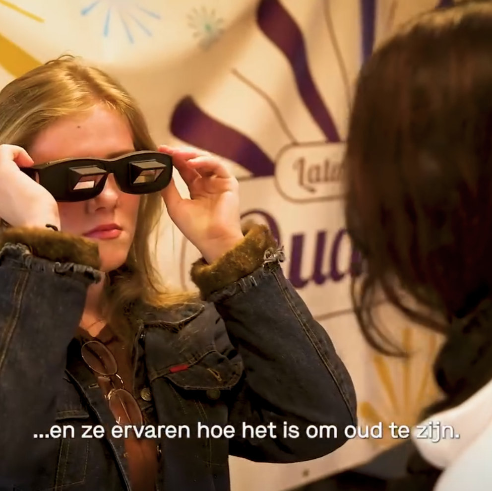 Vrouw die probeert 3D-brillen te passen terwijl ze met iemand anders praat, op de achtergrond een banner met tekst.