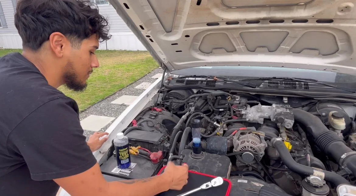 A young man inspecting the engine of a white car with the hood open outdoors in a grassy area.