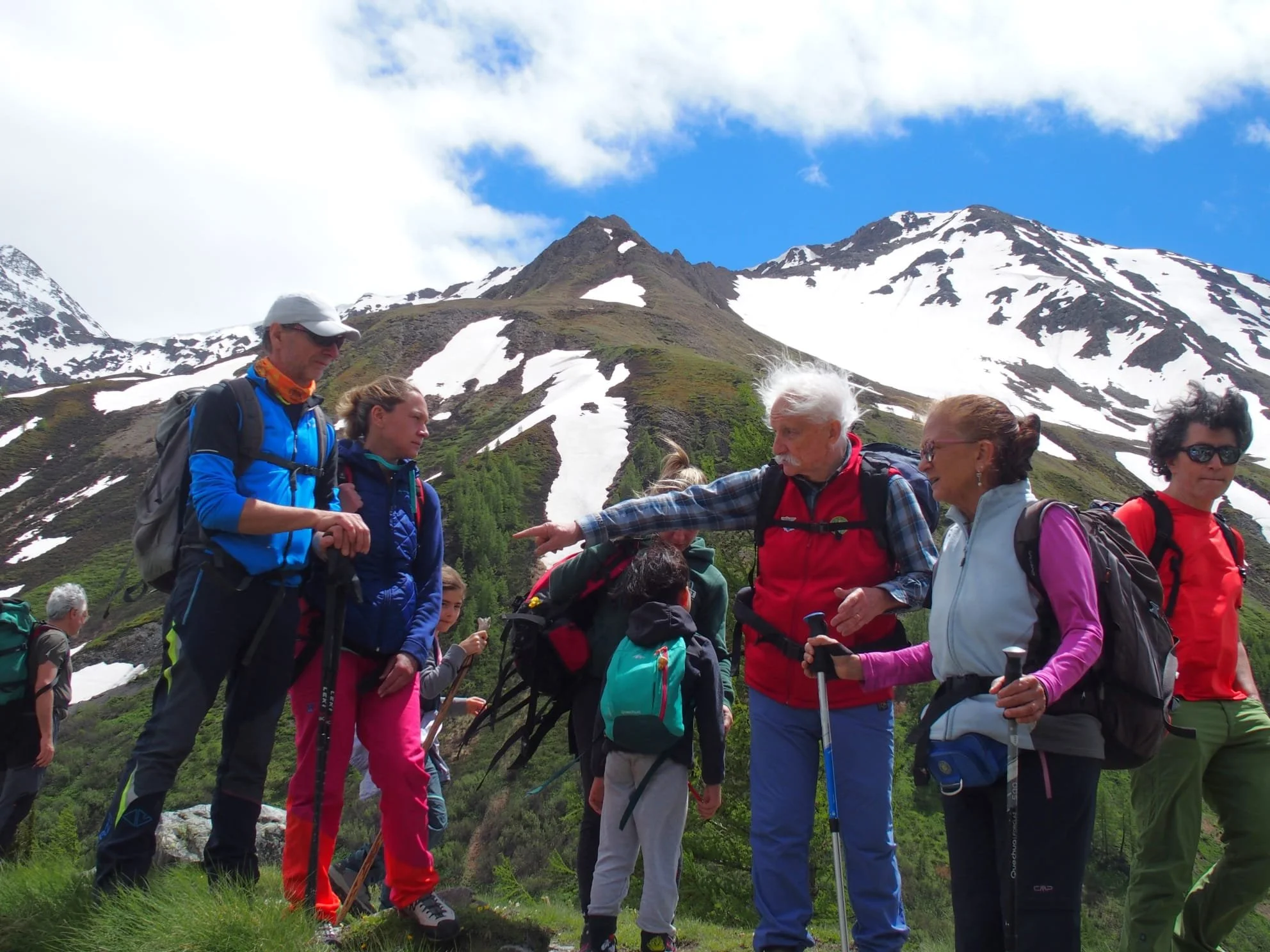 Gruppo di escursionisti in montagna con neve e verdi colline, alcuni che ascoltano un uomo anziano che spiega.