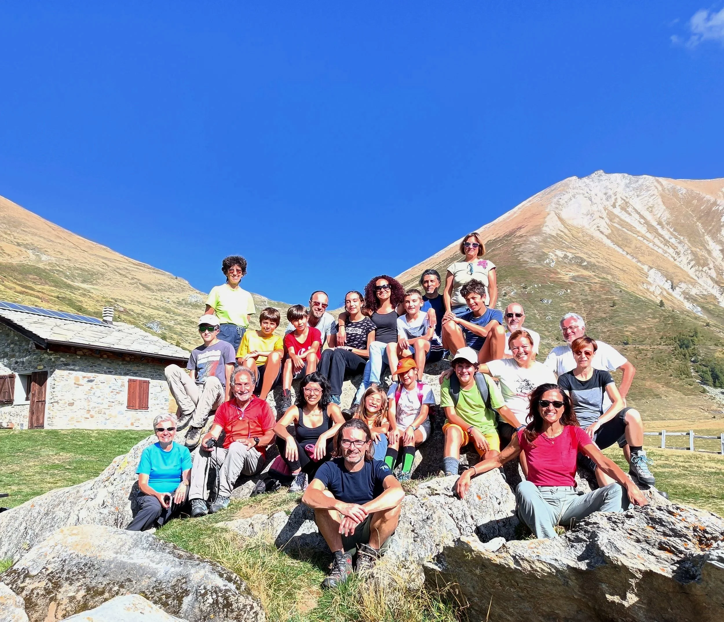 Gruppo di persone su una roccia in montagna sotto un cielo azzurro, alcune con abbigliamento sportivo e occhiali da sole.