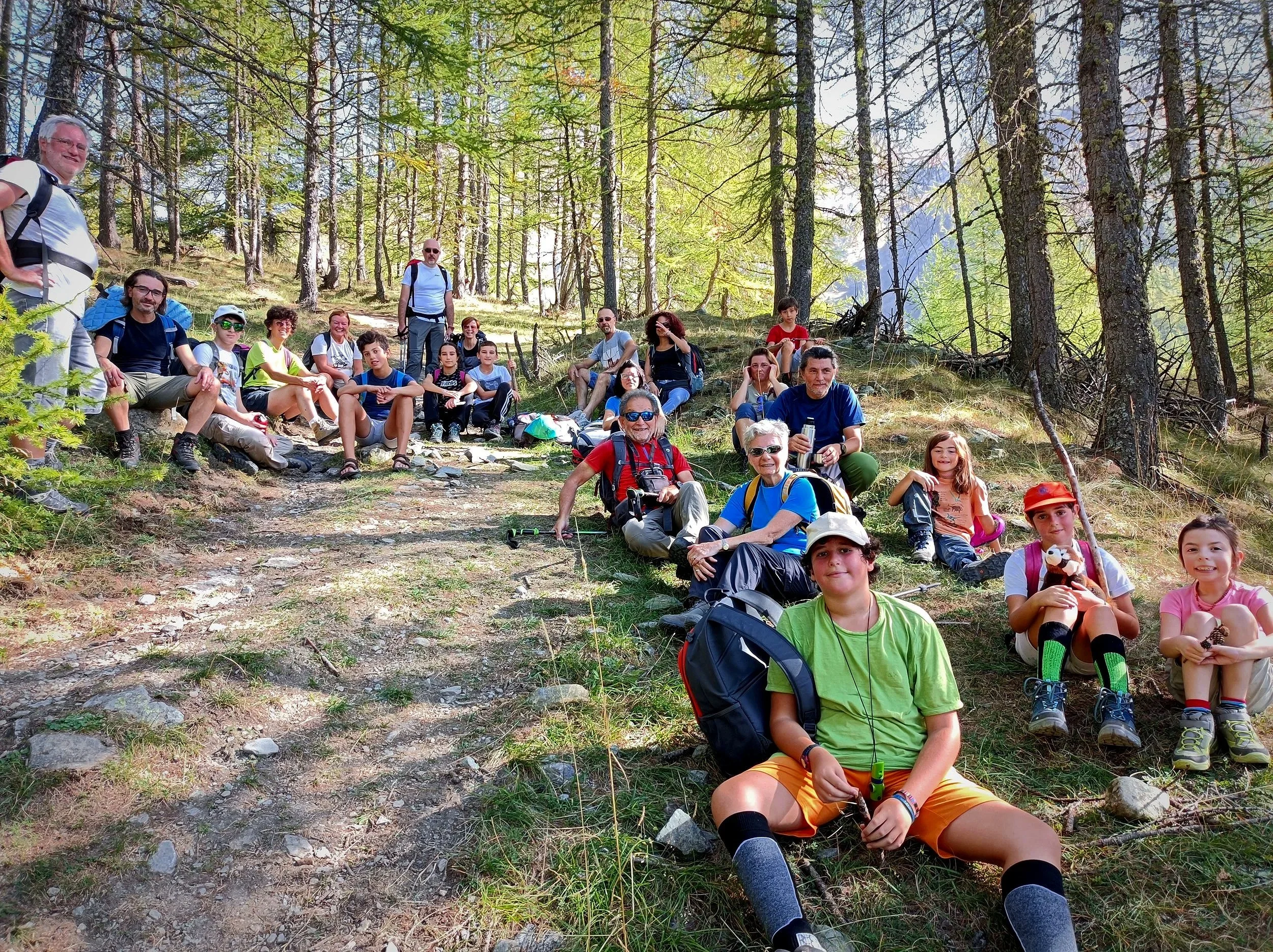 Gruppo di persone di varie età che fa un'escursione in un bosco, seduti e in piedi su un sentiero tra gli alberi.