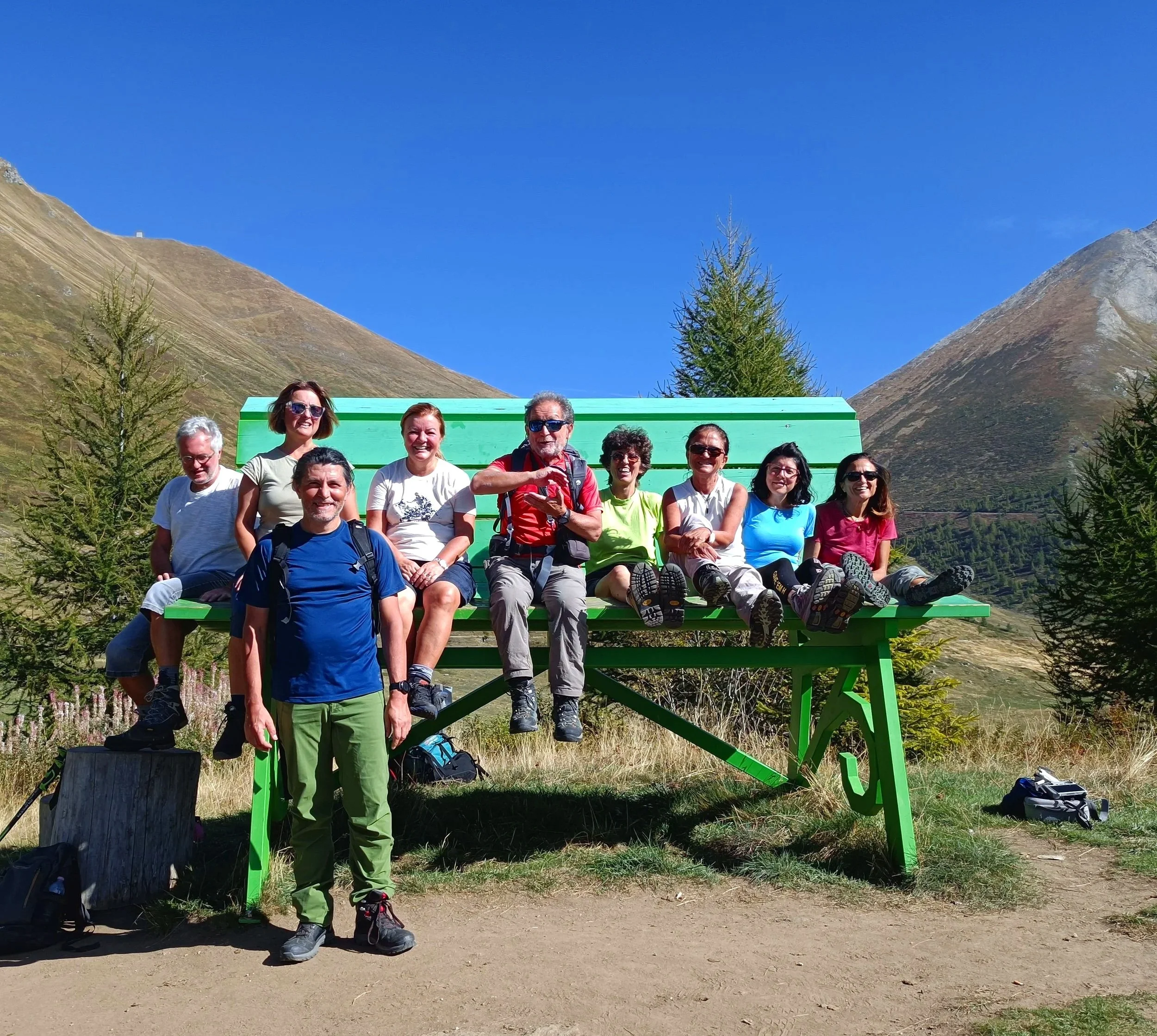 Gruppo di persone che si riposa su una grande panchina verde in un paesaggio montano con cielo blu e pini.