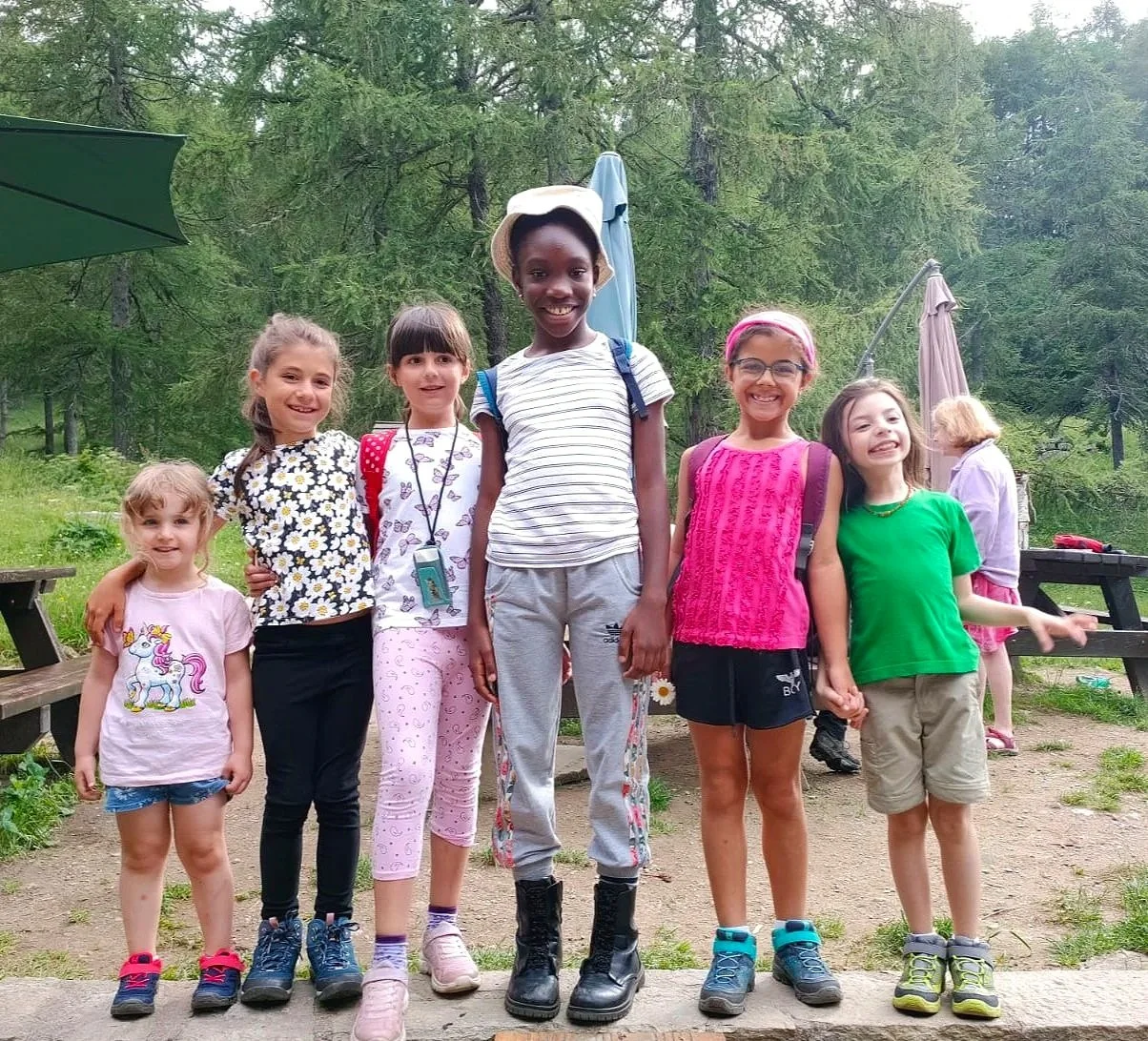 Gruppo di sette bambini sorridenti che posano all'aperto in un'area woodland, alcune con zaini e abiti colorati.
