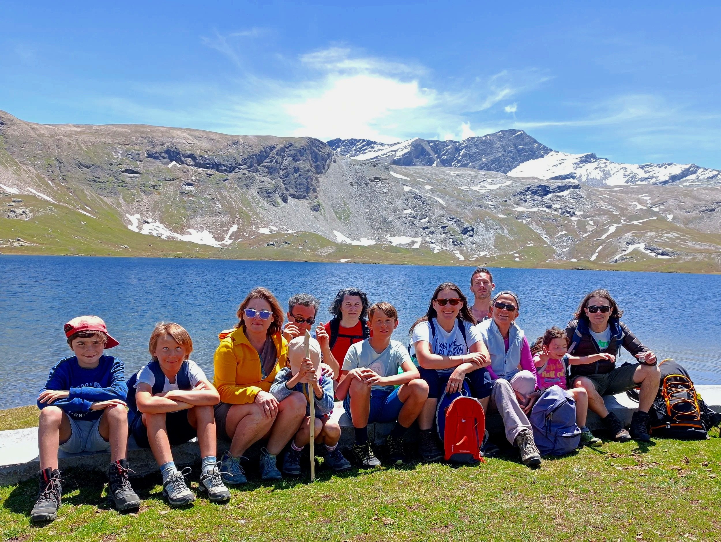 Gruppo di persone sedute su un prato vicino a un lago di montagna, con sfondo di montagne e cielo azzurro.
