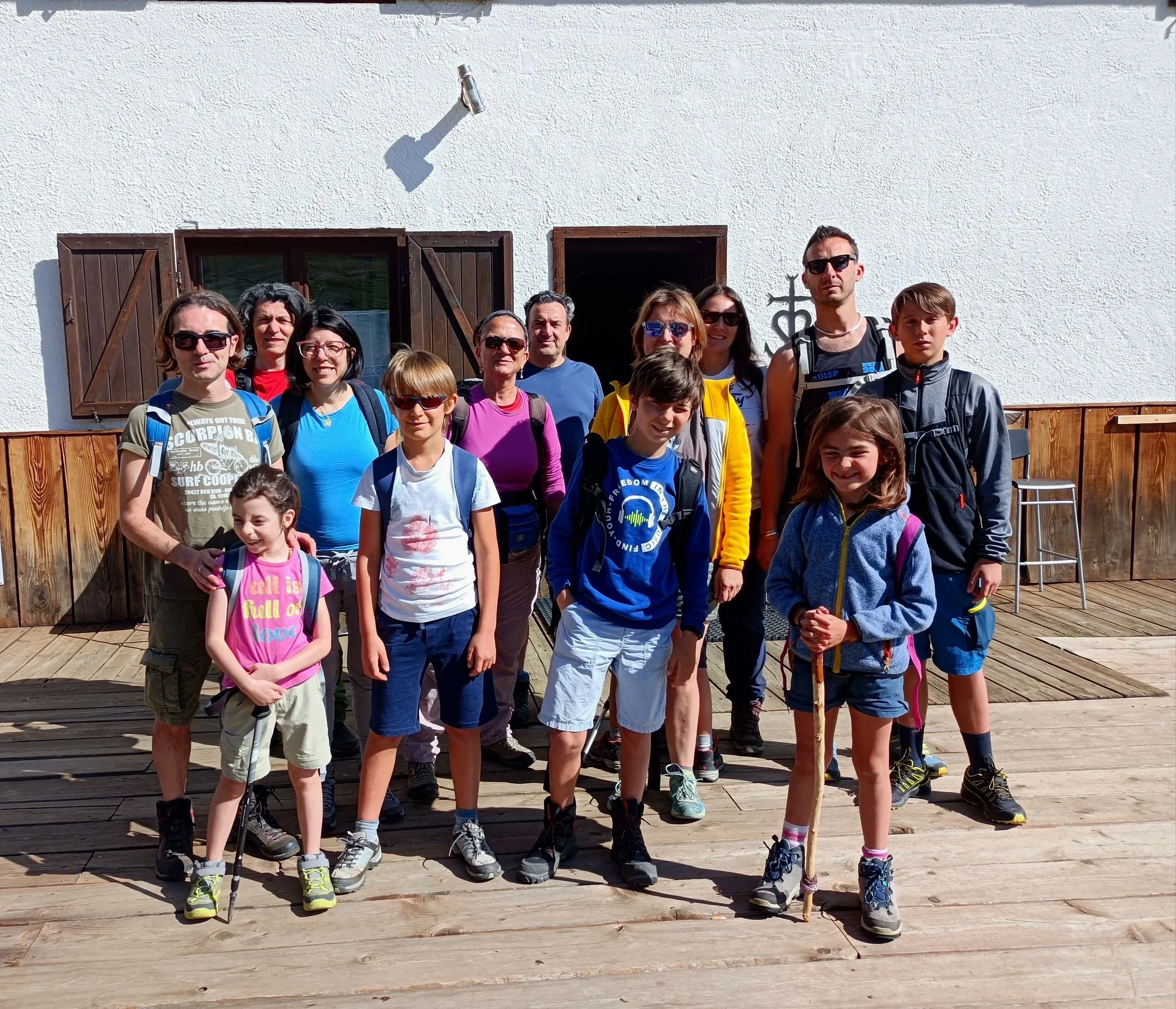 Un gruppo di persone, adulti e bambini, posano per una foto all'aperto in un'area con pavimentazione in legno, con una parete bianca e alcune finestre con persiane di legno di sfondo.