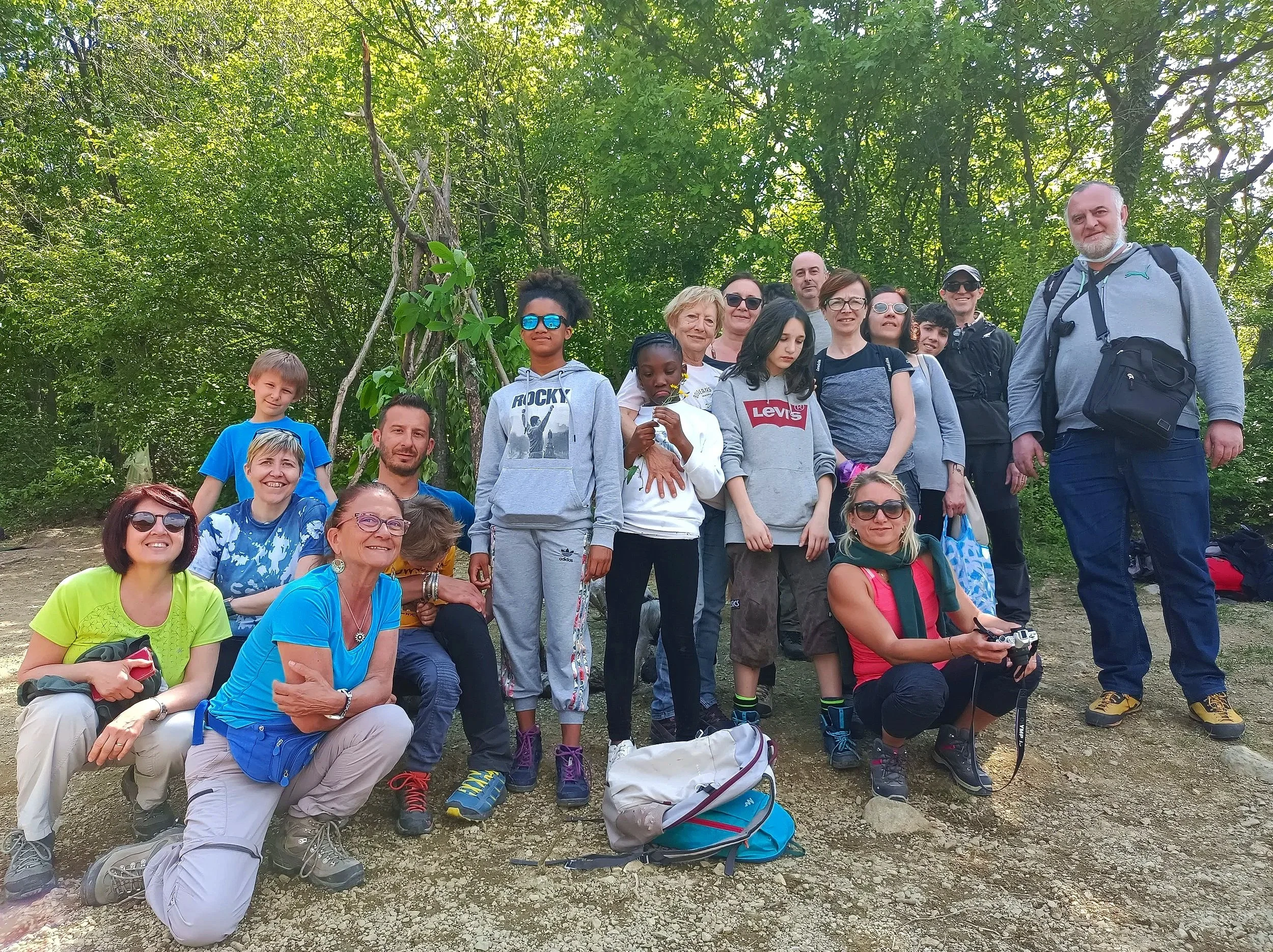 Gruppo di persone che si radunano in un'area boschiva durante un'escursione, alcuni sorridono e indossano abbigliamento casual e da trekking.