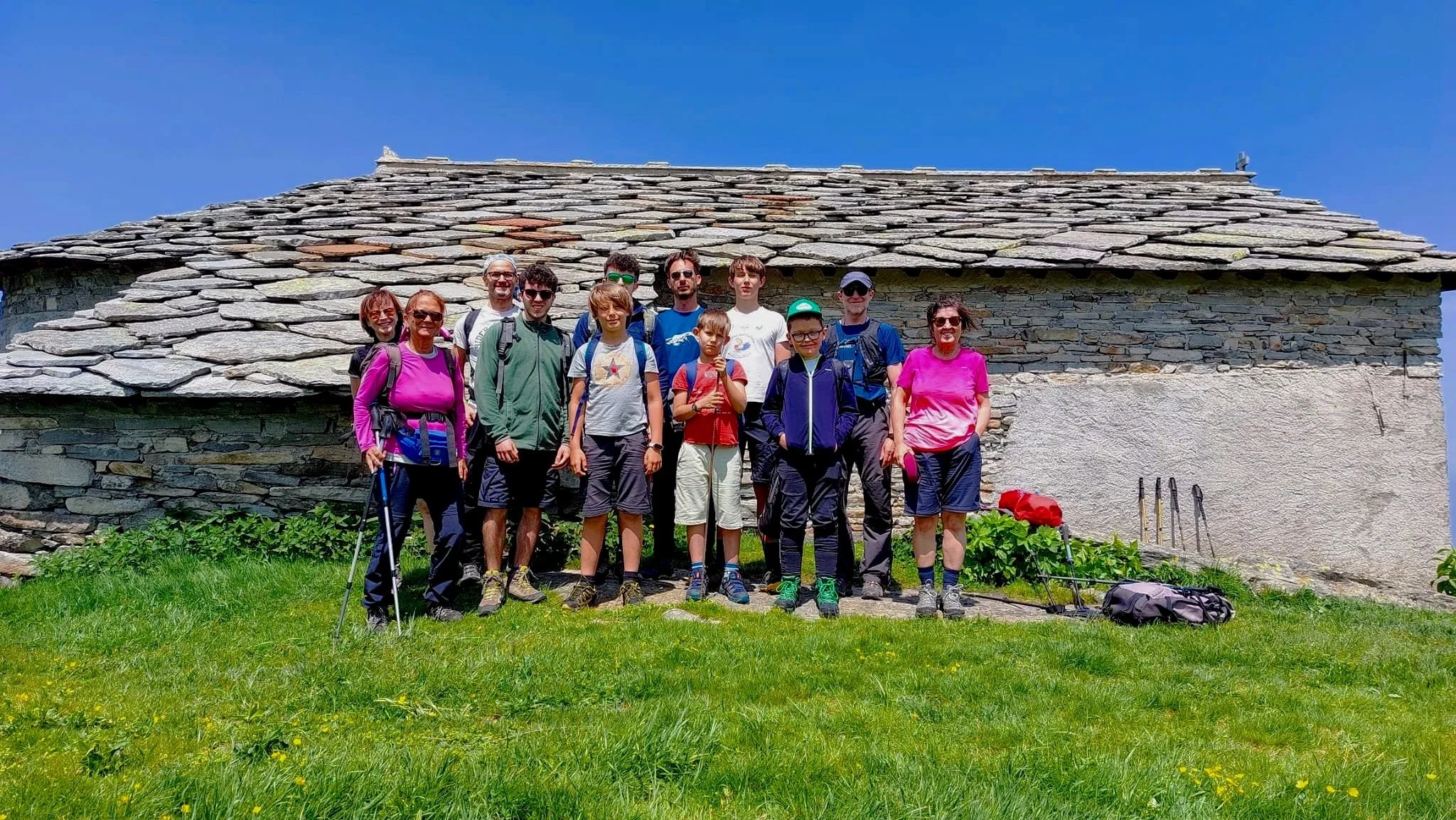 Gruppo di persone che fanno trekking davanti a un edificio in pietra sotto un cielo azzurro.